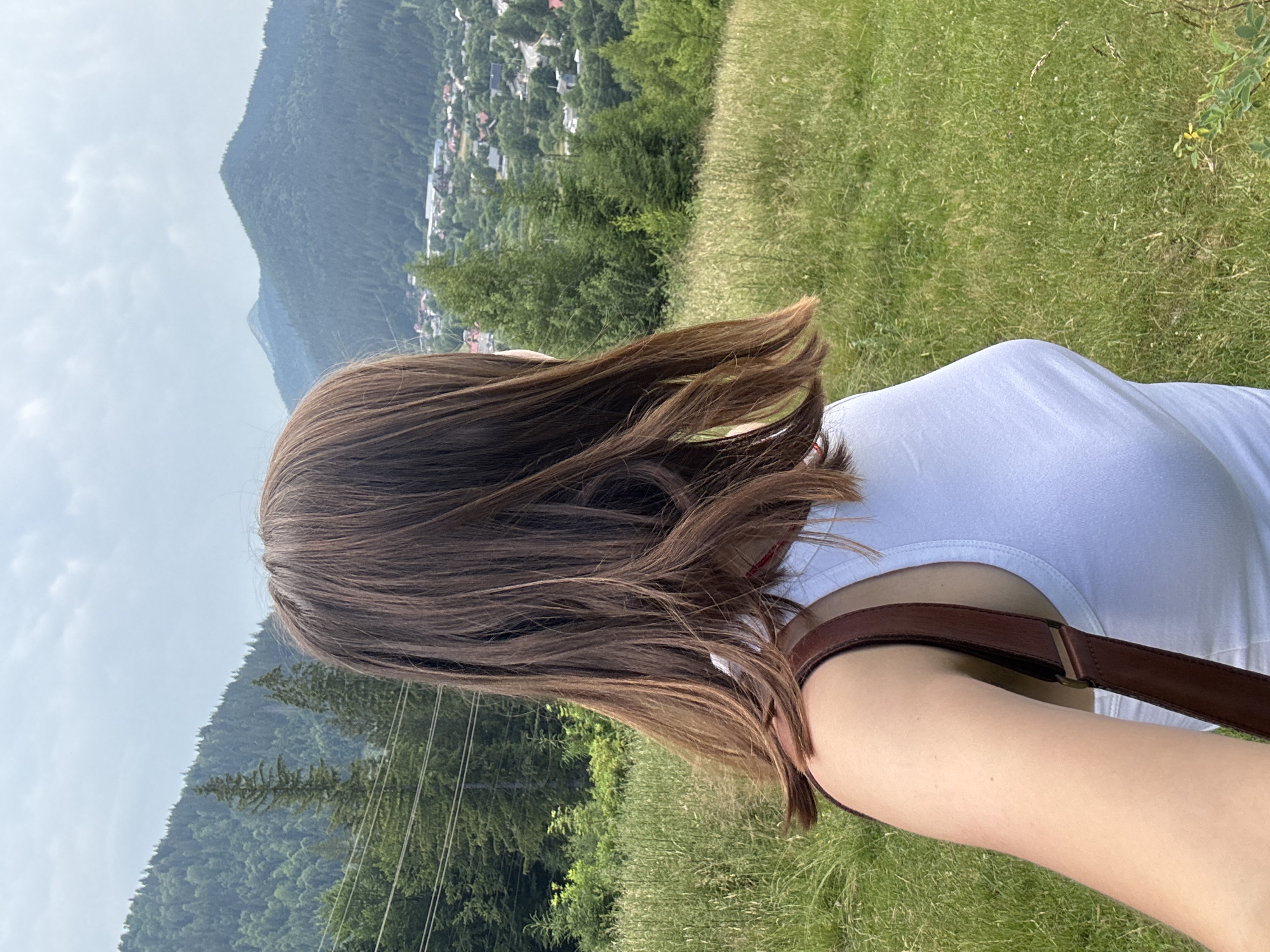 A young woman with brown hair is seen from behind, facing away from the camera, in a scenic outdoor location. She is wearing a white tank top and a brown strap from a bag rests on her shoulder. The background consists of a lush green grassy hill, dense evergreen trees, and a distant view of a town nestled among rolling mountains under a cloudy sky. The image captures a peaceful moment in a natural setting. The location is identified as Tatariv, Ukraine.