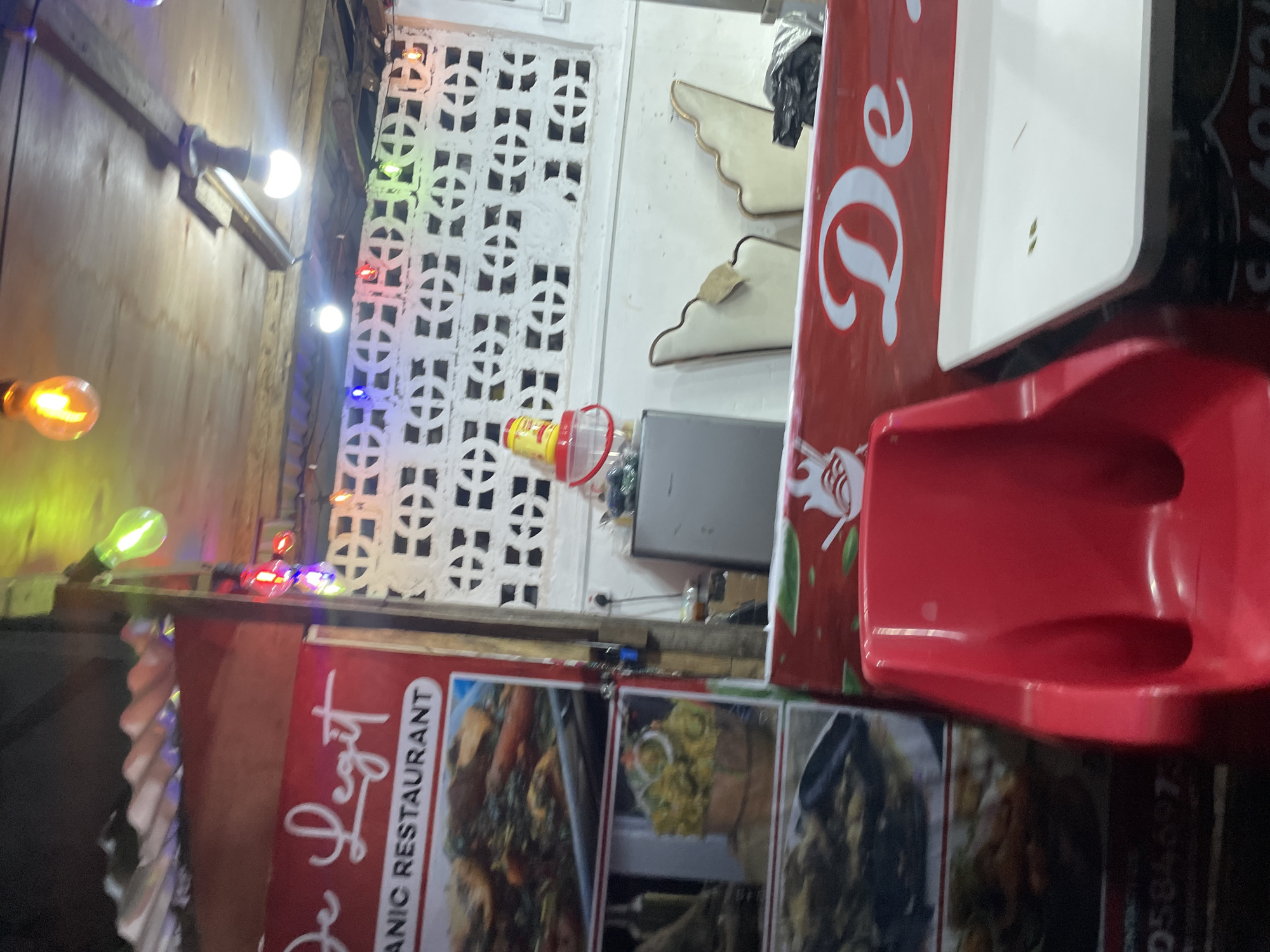 The image depicts a street-side restaurant in Ado, Nigeria. A red sign with white cursive lettering that reads "De Legit" and "ORGANIC RESTAURANT" is prominently displayed. Below the sign are photographic displays of various food dishes. To the right of the sign is a white patterned wall. String lights with colorful bulbs are draped across the ceiling, contributing to the atmosphere. In the foreground, a bright red plastic chair is visible, and to its right, a white table surface. A small grey refrigerator is also present behind the counter area. The overall impression is of a casual, local eatery.