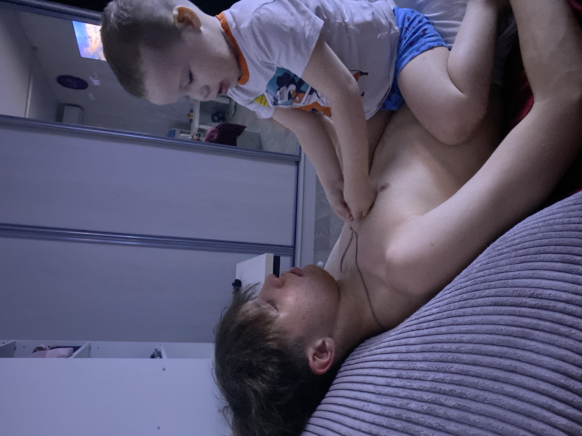 A young child straddles an adult lying on a bed. The child wears a white t-shirt with a colorful graphic and blue shorts. The adult is shirtless and wearing a silver chain around their neck. They are lying on a gray, corduroy-patterned pillow. Behind them, a closet with sliding mirrored doors is visible, reflecting a television screen. A white storage unit with items on its shelves is also present. The location is unidentified, and there is no indication of the city of Sochi, Russia, in this image.