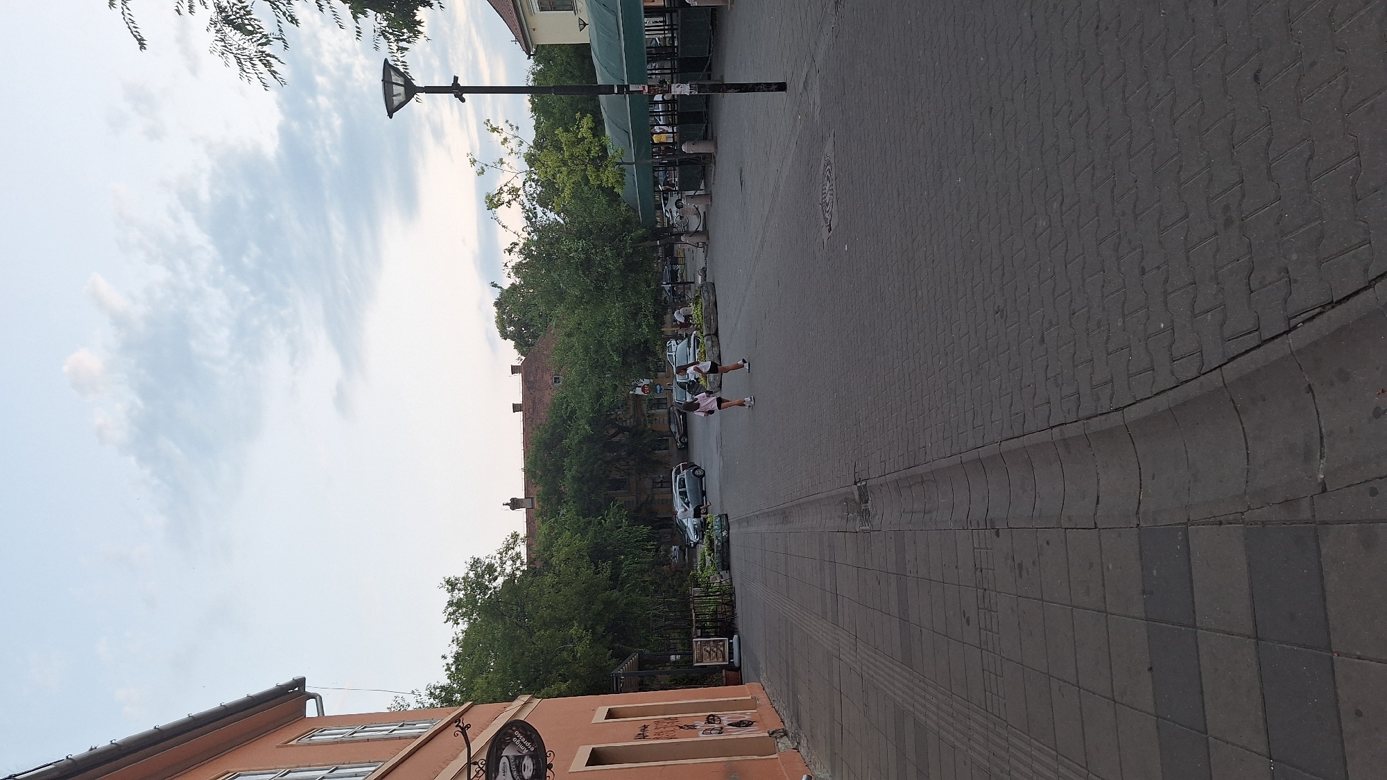 The image depicts a street scene in Sombor, Serbia. In the foreground, a paved sidewalk transitions to a cobblestone street. Two young females are walking on the cobblestone area. To the left, a building with an orange facade and windows is visible. Beyond the street, trees line the background, with several cars parked and a covered outdoor seating area, likely for a cafe or restaurant. A lamppost stands on the right side of the scene. The sky above is partly cloudy.