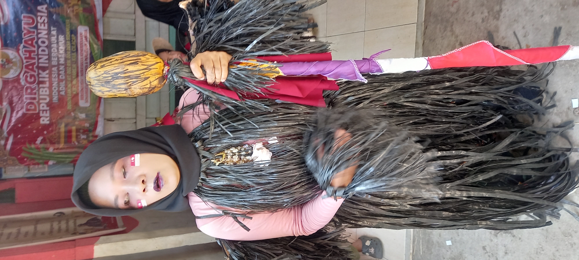 A full-body shot captures a person in a unique costume, standing outdoors in what appears to be the city of Sidamulya, Indonesia. The backdrop features a banner with text in Indonesian, including "DIRGAHAYU REPUBLIK INDONESIA," indicating a celebration of Indonesian Independence Day. The person is wearing a black hijab and a pink long-sleeved shirt. Their costume consists of a dark, layered skirt made of what looks like shredded black material, giving a fringed or feathery appearance. They also have dark, wing-like appendages attached to their shoulders and arms, also made from the same shredded material. A large, golden pendant with multiple beads and white accents hangs from their neck. In their hands, the person holds a tall staff adorned with colorful fabric strips in shades of red, purple, and white, culminating in a bulbous, yellowish object at the top. The person's face is partially visible, with white and red face paint applied to their cheeks, resembling the Indonesian flag. Dark lipstick completes the makeup. In the background, other individuals are partially visible, including one wearing a hat and another in a black hijab. A child's foot wearing a grey shoe is visible on the left side of the frame. The ground is a tiled surface.