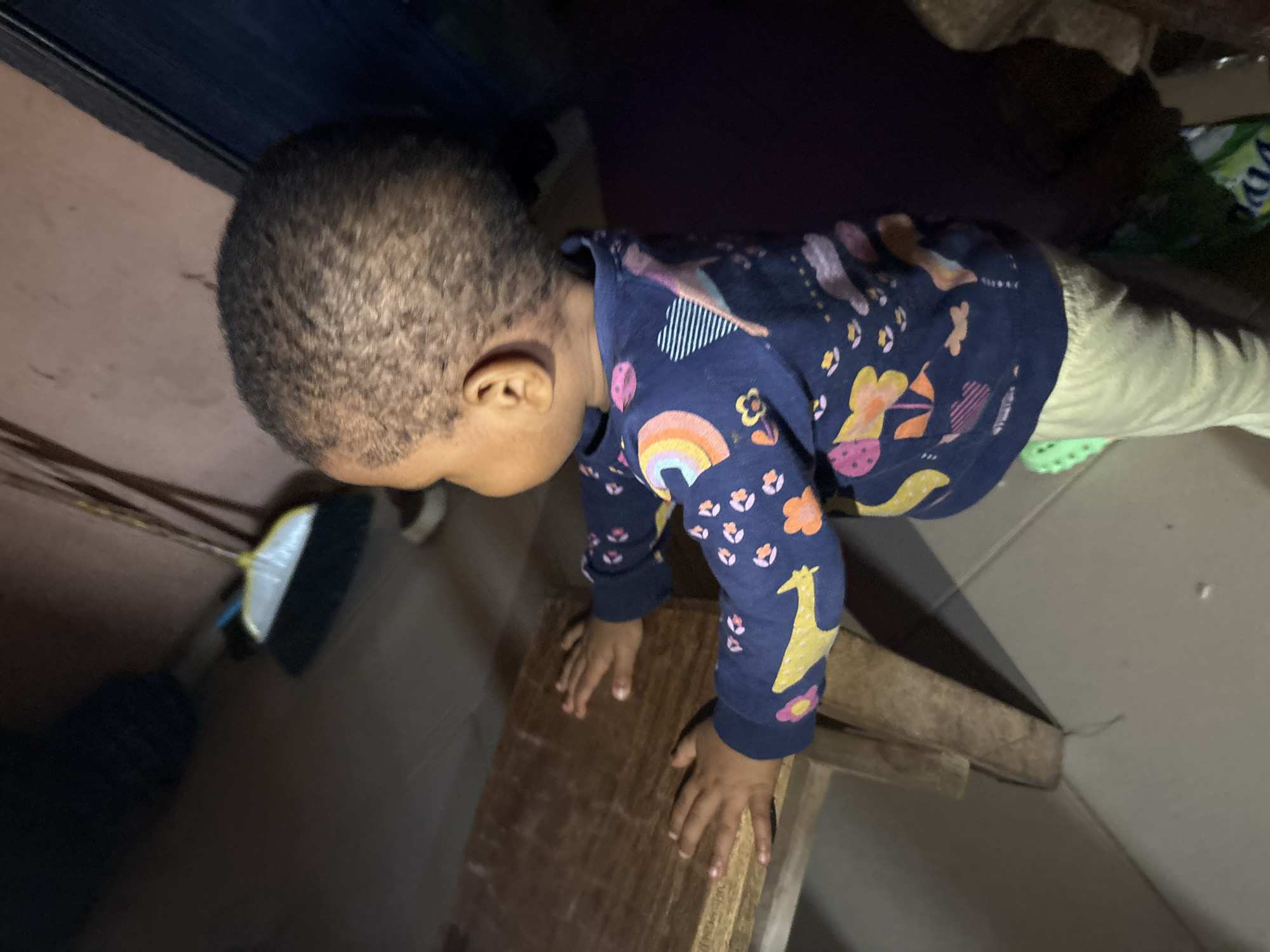 A toddler, facing away from the camera, stands and leans on a wooden table. The toddler wears a dark blue long-sleeved shirt with a colorful pattern of abstract shapes and animals. Light-colored pants are visible at the waist and legs. The toddler's hair is short and dark. The toddler's hands are placed on the surface of the wooden table, which appears to be unfinished or worn. The background is dimly lit and shows a tiled floor and a broom leaning against a wall. The scene suggests an indoor environment, possibly a home, in Onitsha, Nigeria.