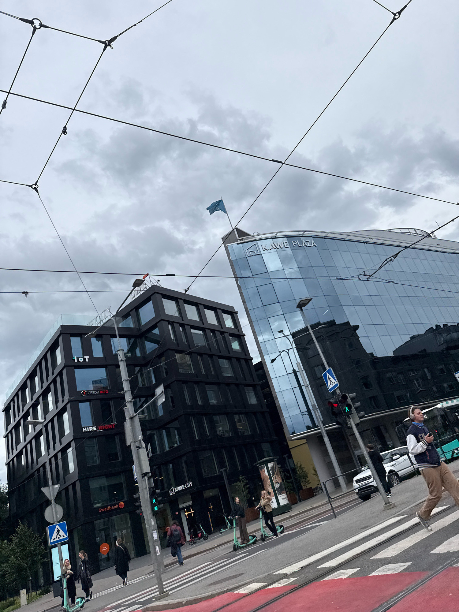 This image captures a street scene in Tallinn, Estonia, likely near Kawe Plaza, a modern glass-fronted building. The sky is overcast with heavy clouds, and overhead, a network of tram or trolleybus wires crisscrosses the frame.
In the foreground, a crosswalk with red markings is visible. Several people are crossing the street. One person, wearing headphones and a varsity-style jacket, is walking towards the right side of the frame. To the left, three individuals are standing near electric scooters, two of whom appear to be on them.
Two prominent buildings dominate the background. To the left is a dark, multi-story building with numerous windows and prominent signage including "1oT", "HIRE RIGHT", and "Swedbank". To the right, the contemporary glass facade of "KAWE PLAZA" reflects the surrounding environment and sky. A blue flag flies from a pole atop one of the buildings.
Traffic lights and street signs are present. A traffic light shows a green signal. A white SUV is parked on the right side of the street, and a green bus or trolley is partially visible further down. The overall impression is of an urban environment with active pedestrian and public transport elements under a cloudy sky.