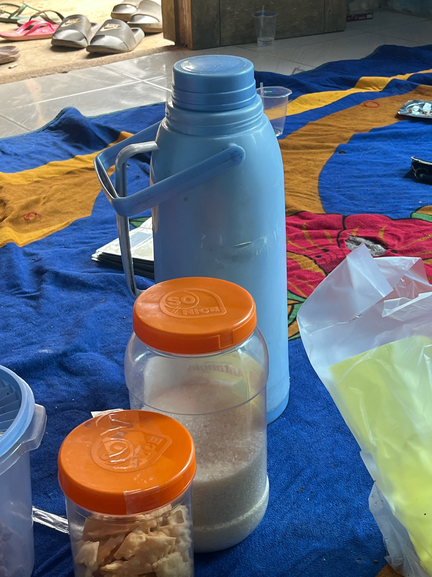 The image depicts an outdoor scene, likely a courtyard or patio, in Kebumen, Indonesia. In the foreground, a blue and yellow striped blanket is spread on the ground. On the blanket, there are two clear plastic jars with orange lids, one filled with white granular sugar and another with what appears to be a snack food. A large light blue thermos is also present, along with a translucent plastic bag containing a yellow item. In the background, several pairs of flip-flops and sandals are visible near an entrance, suggesting a casual domestic setting. The overall impression is that of an informal gathering or a moment of relaxation outdoors.