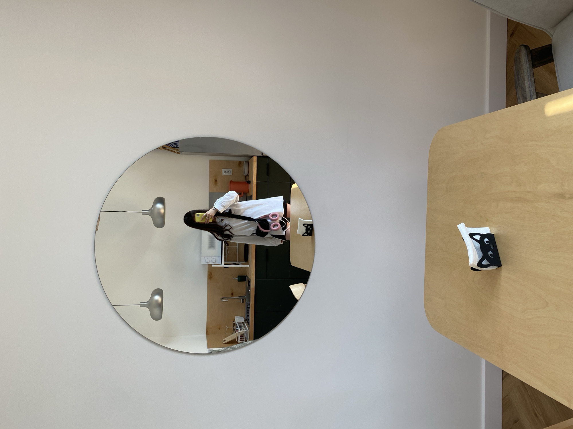 The image shows a portion of a room with a circular mirror mounted on a light-colored wall. The mirror reflects a person holding a yellow phone, seemingly taking a selfie. The person is wearing a white top, black shorts, and a pink fanny pack. Behind the person in the reflection is what appears to be a kitchen area with cabinets and a sink. To the right of the mirror, there's a light wooden table with a black and white tissue holder. The location is not discernible from the visual content.
