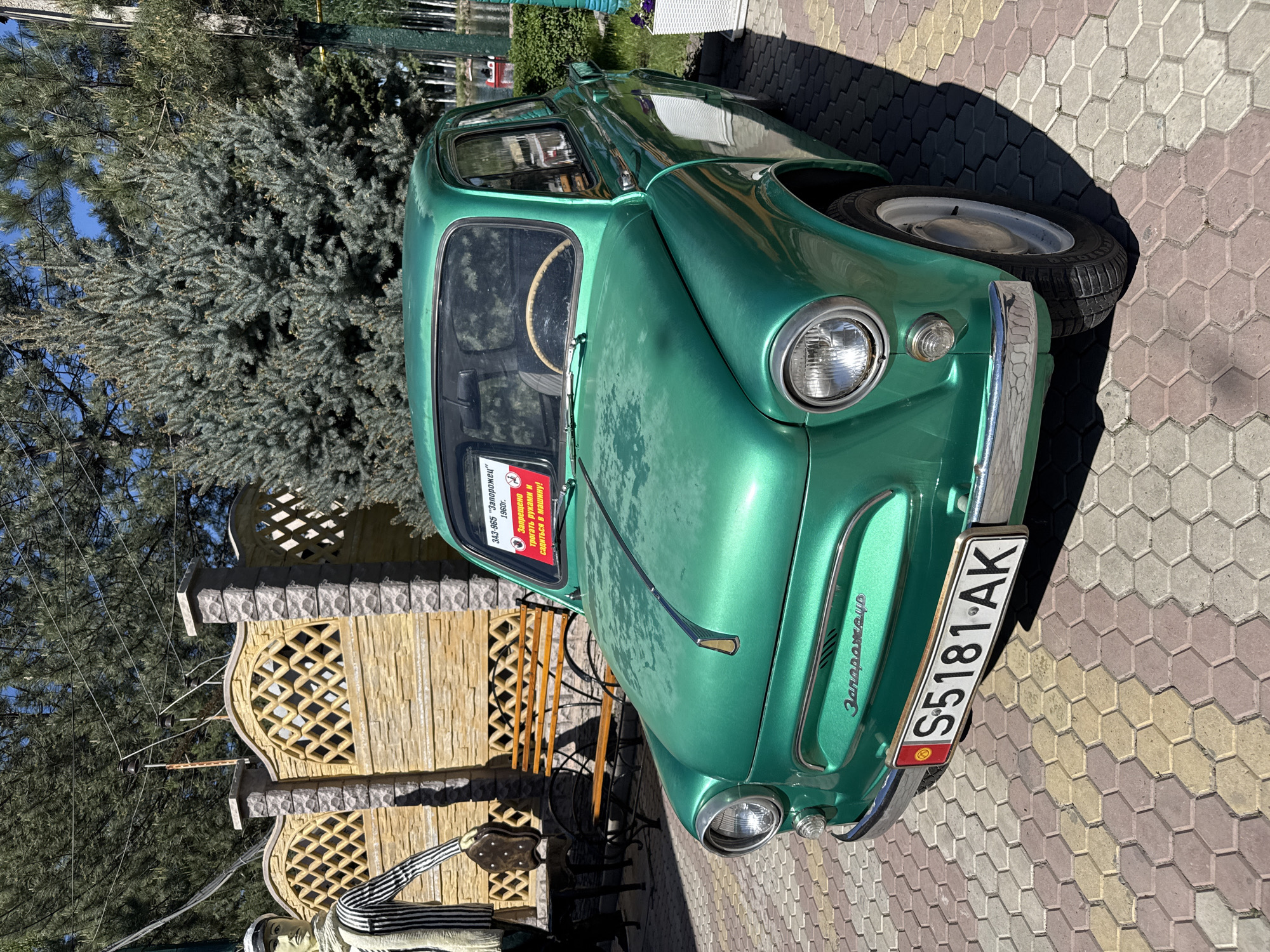 The image shows a green vintage car, identified as a ZAZ-965 "Zaporozhets" from 1960. The car is located in Kenbulun, Kyrgyzstan, indicated by the license plate reading "S5181-AK," with the Kyrgyz flag displayed. The car is parked on a hexagonal-paved surface next to a wooden decorative fence and a tree. A sign is attached to the car’s windshield, stating in Russian, "Запрещено трогать руками и садиться в машину," meaning "It is forbidden to touch and sit in the car."