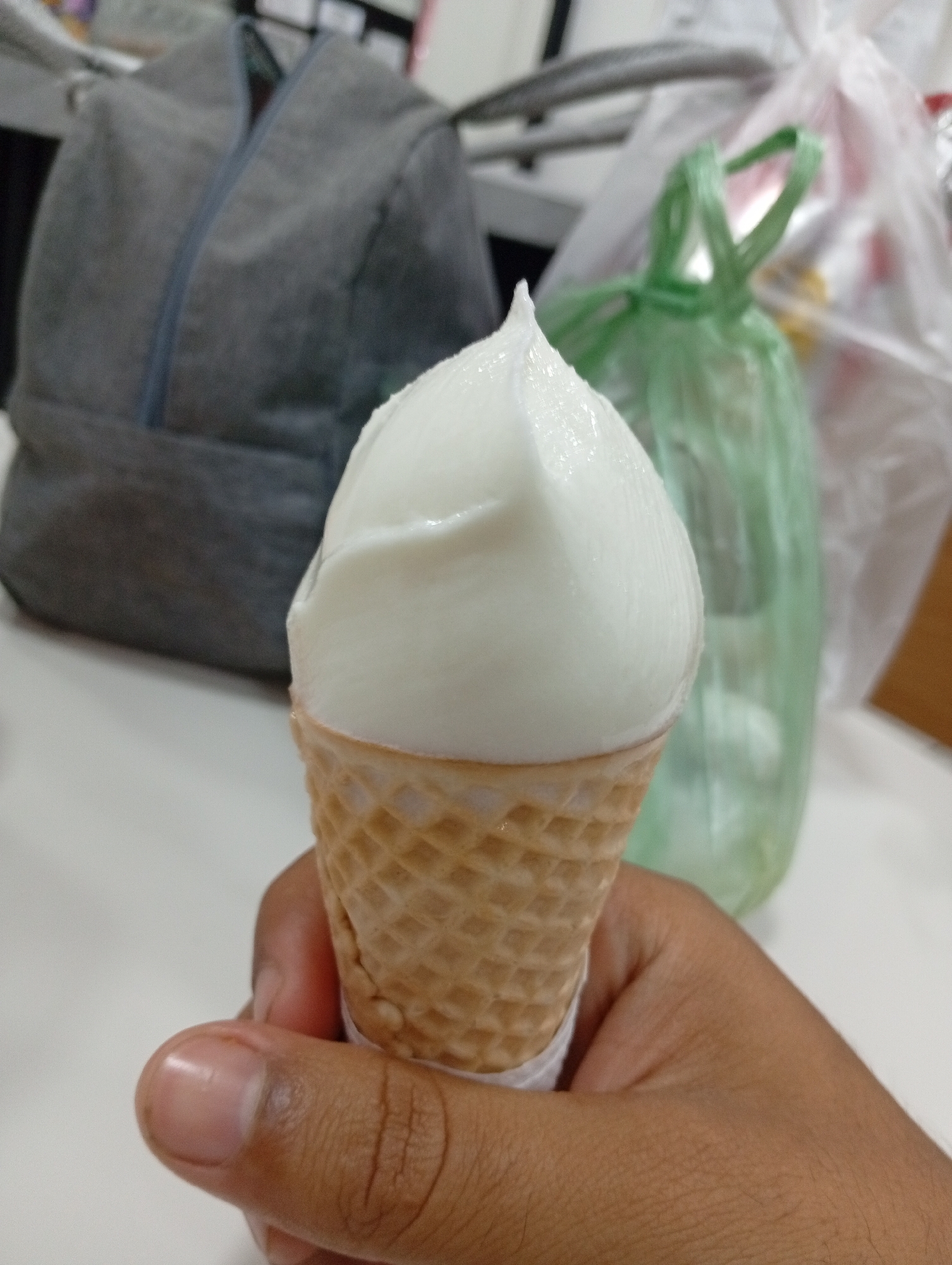 A hand holds a small ice cream cone with white ice cream in front of a white table. Behind the ice cream are a gray zippered bag and a green plastic bag. The location is not specified. The scene contains no information related to Juan Griego, Venezuela.