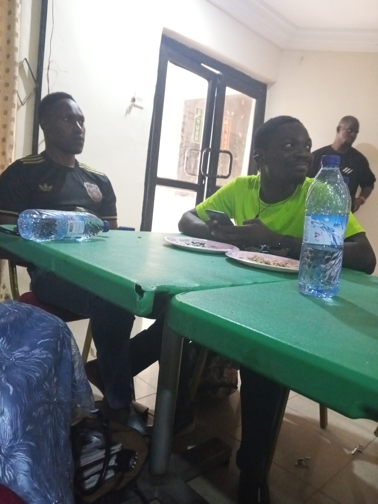 The image shows three men in an indoor setting. Two are seated at a green table with two plates and two water bottles. One man is wearing a black shirt and the other a green shirt. The man in the green shirt appears to be using a phone. A third man, wearing a black shirt, is standing in the background near a doorway with a glass door. A blue chair is partially visible at the bottom left. The location is an unknown city and region.