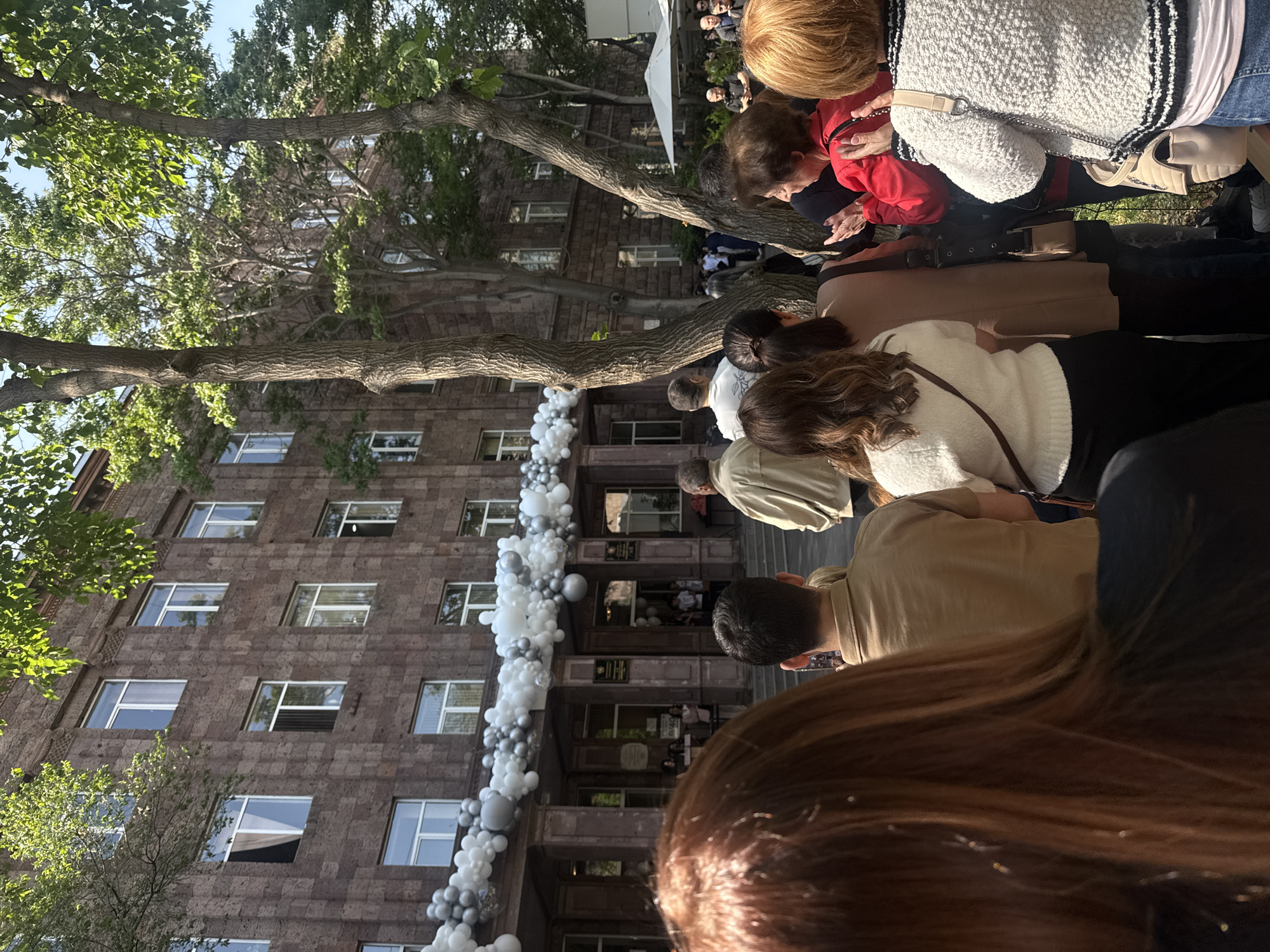 The image captures a scene in Yerevan, Armenia, likely in front of a significant building due to the presence of decorations, people, and the building itself. The building, constructed from a dark stone, features multiple windows across its several stories. The entrance is adorned with a decorative balloon garland in silver and white colors.
Several individuals are visible in the foreground and extending toward the building. People are dressed in casual clothing. Many are turned toward the building, possibly observing an event or activity taking place there.
The environment includes trees with green leaves, partially obscuring the building. The overall scene suggests an event or gathering.