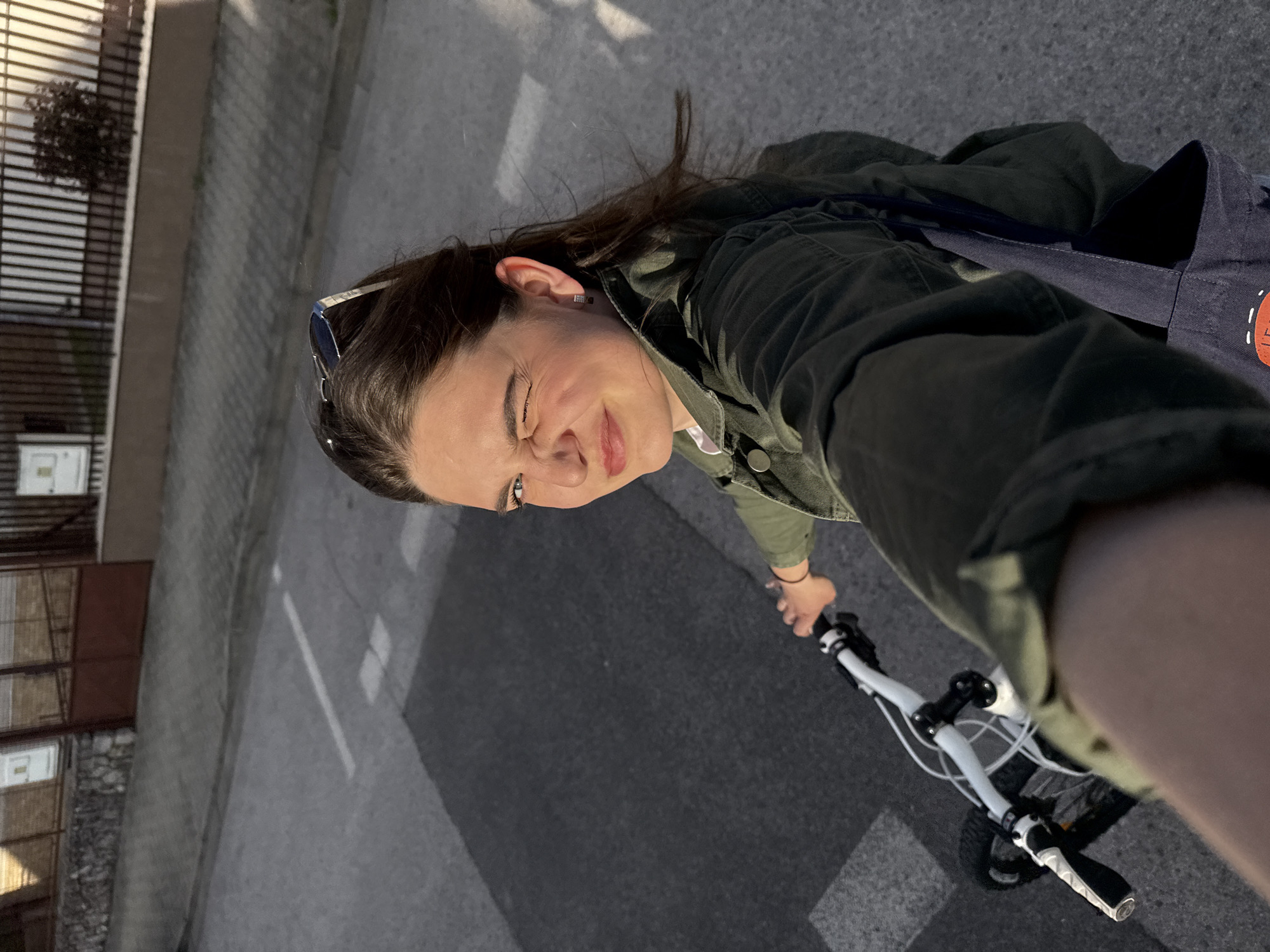 A young woman is riding a bicycle on a street in Trnava, Slovakia. She is wearing an olive green jacket and has sunglasses perched on her head. She is also wearing a dark-colored tote bag over her shoulder. The road is paved with asphalt and has white lines marking lanes. Buildings line the side of the street, showing the urban environment. The time of day appears to be late afternoon.