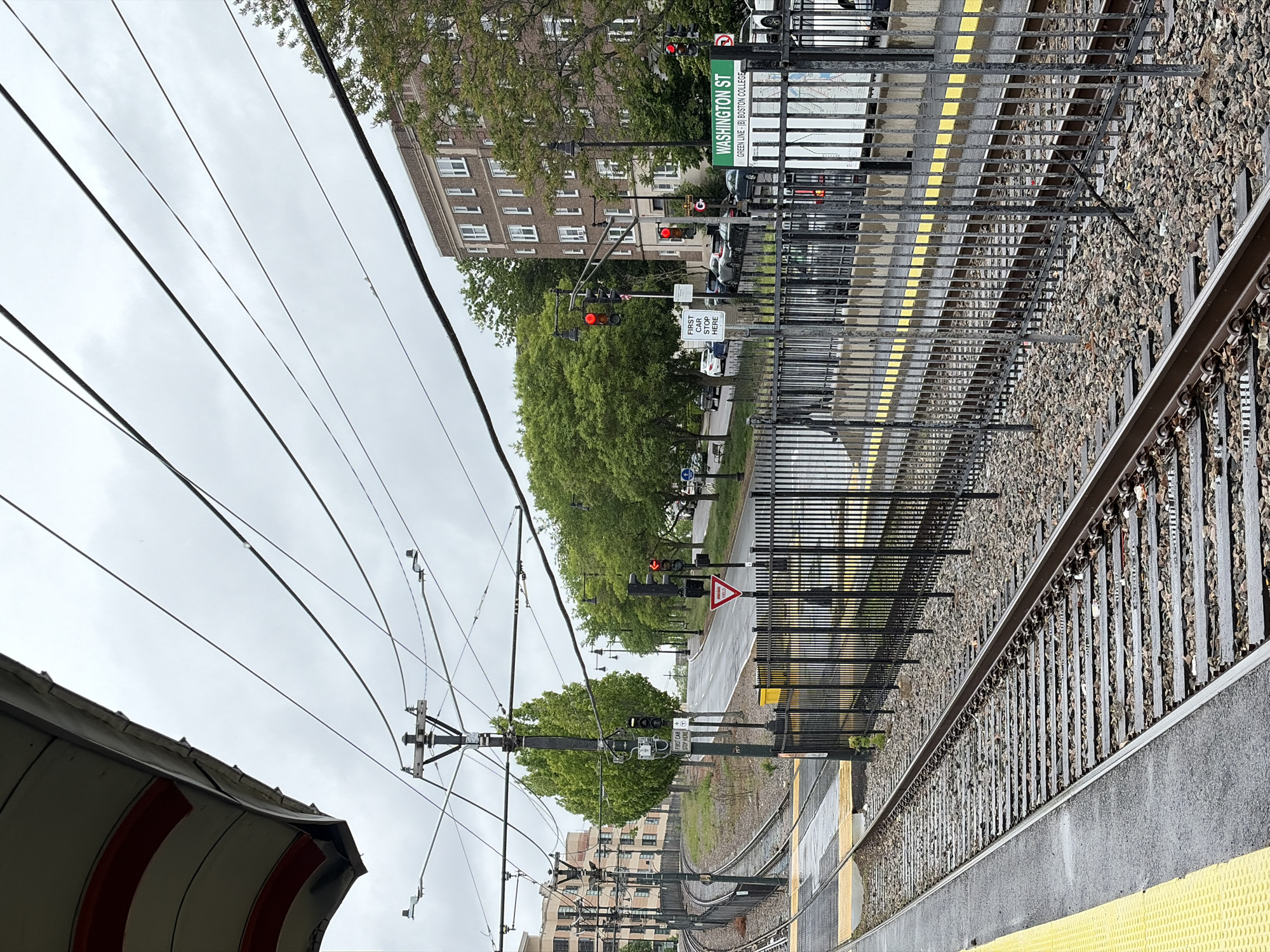 This image depicts the "Washington Street" Green Line station located on the "B" branch in Boston, Massachusetts. The perspective is from the inbound platform facing towards the south and west. The station platform is edged with a yellow safety strip and lined with a black metal fence separating the platform from the tracks. Beyond the fence are the tracks and the ballast bed. Overhead are the electric catenary lines supporting the Green Line streetcars. Street-level traffic lights are visible beyond the tracks. The station sign identifies the location as "Washington Street" and the "Green Line, (B) Boston College." A building is visible behind the station, and the sky is overcast. A sign is visible which indicates the location of where the first car stops.