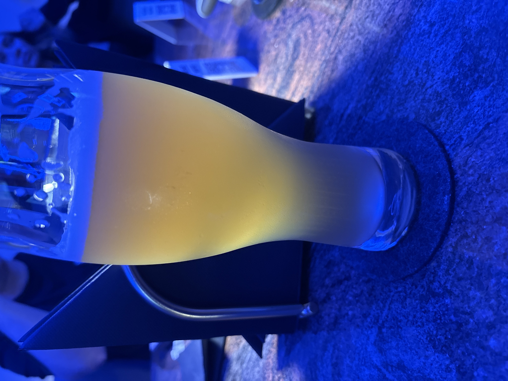 A tall, frosted glass filled with a pale yellow, frothy beverage, likely beer, sits on a dark, textured surface bathed in blue light, suggesting a bar or restaurant setting in Budva, Montenegro. A black napkin holder with a silver rim is partially visible behind the glass. In the blurred background, two figures can be seen, one wearing a dark top, and another figure with lighter hair. The overall ambiance is dim and atmospheric, with the blue lighting creating a moody scene.