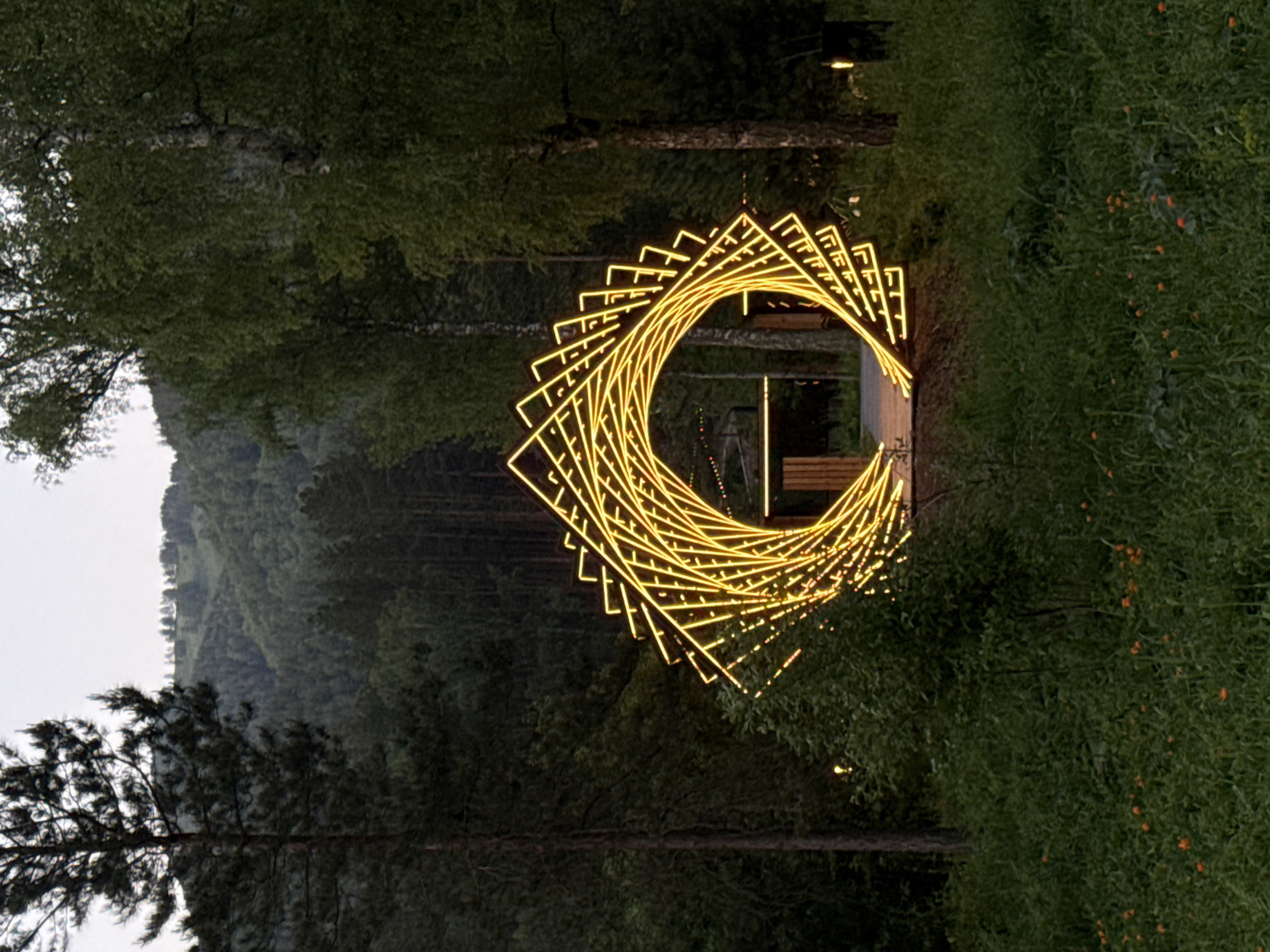 The image displays an illuminated art installation set within a natural, forested landscape at dusk or early night in Ust'-Muny, Russia. The central feature is a large, glowing archway or tunnel, constructed from numerous golden-yellow light strips that form a series of twisting, rectangular frames. This design creates a dynamic, spiraling effect that serves as an entrance or pathway.
The archway is flanked by dense vegetation. On the right, lush green bushes and foliage are visible, interspersed with small orange flowers or berries. To the left, several large trees with dark green canopies, including both coniferous and deciduous types, frame the scene. In the background, rolling hills or mountains covered with thick forests recede into the distance, meeting a light grey sky.
Through the illuminated arch, faint lights from what appears to be a continuation of a path or another lighted area can be discerned. A wooden structure, possibly a bench or part of a platform, is also subtly visible within the glowing tunnel. The time of day is clearly indicated by the dim natural light and the prominent artificial illumination of the art piece. The scene conveys a tranquil and enchanting ambiance, blending natural elements with modern artistic design. No people or visible text are present in the image.