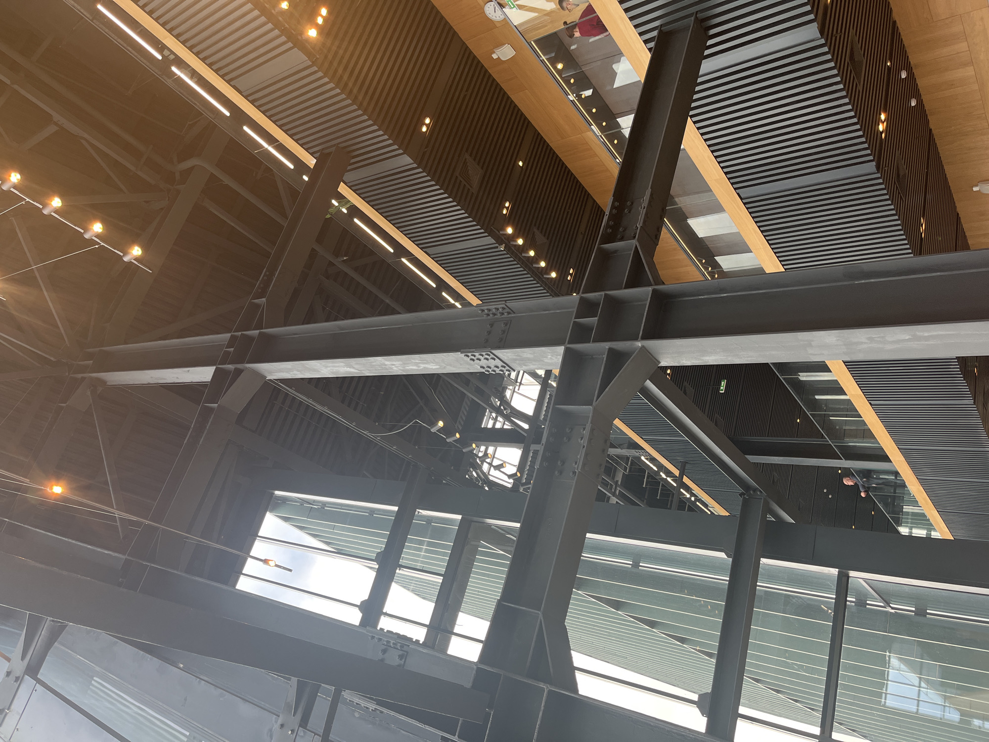 Captured from a low angle, the image showcases the complex architecture of an indoor space. The primary elements are black steel beams configured in an elaborate structural support system, with numerous bolts visible, indicating sturdy construction. Overhead, a latticed ceiling is composed of alternating panels of wood and dark, possibly metal, slats. Further up, a glassed-in area gives a glimpse of what appears to be an upper level, where a person can be partially seen. The environment is well-lit, likely by both artificial sources and natural light filtering through windows. The scene is industrial or commercial, suggesting a modern building design. The city and specific location are not discernible from the image.
