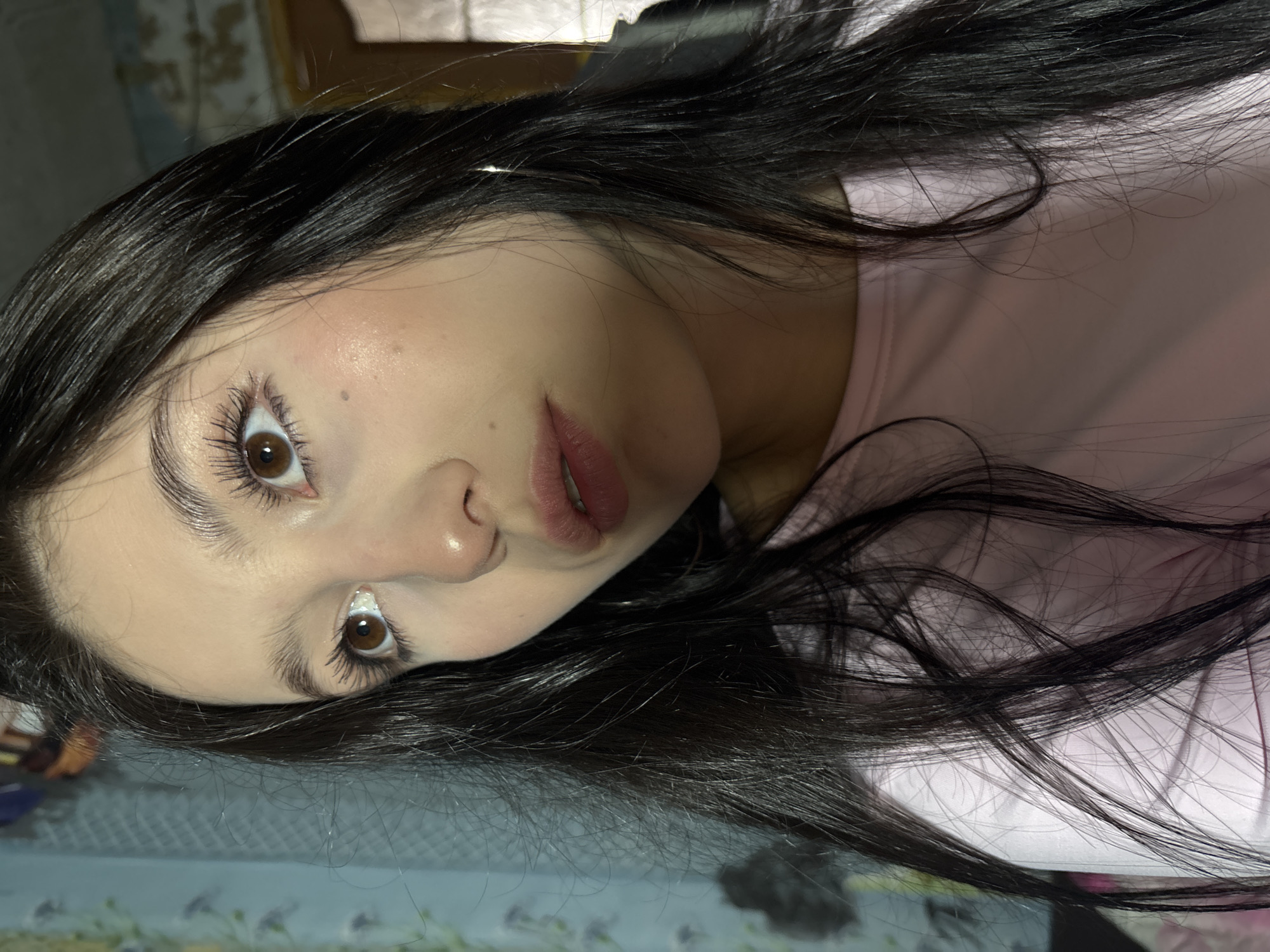 An indoor, eye-level portrait depicts a young woman in Фоки, Russia. She has long, dark hair, brown eyes, and fair skin, wearing a plain light pink long-sleeved shirt. Her gaze is directed upwards and slightly to her left. Her lips are slightly parted, and a faint mole is visible on her right cheek. A thin hoop earring is present on her right ear. The background, out of focus, consists of a wall covered in light blue or teal patterned wallpaper, with visible signs of peeling and discoloration in the upper right area.