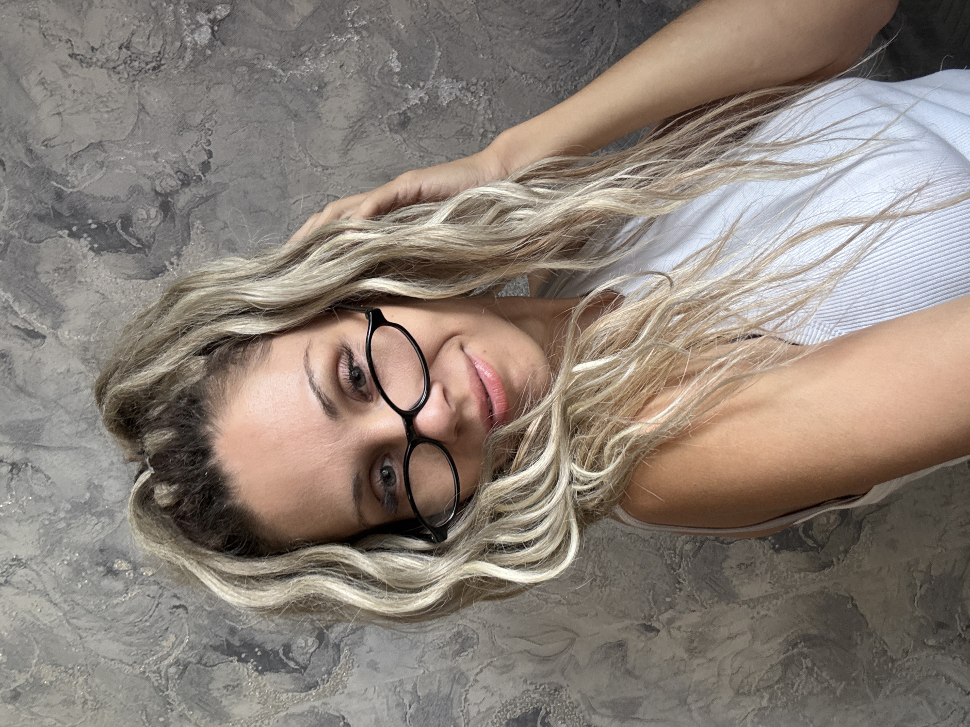 The image shows a medium shot of a woman with blonde, wavy hair pulled up in a high ponytail, wearing black-rimmed glasses. She is looking slightly to her right, with a subtle smile. Her right hand is raised and touching her hair. She is wearing a white, ribbed tank top. The background is a textured, grey wall. The image does not contain any information to suggest it was taken in Rusanovo, Russia.