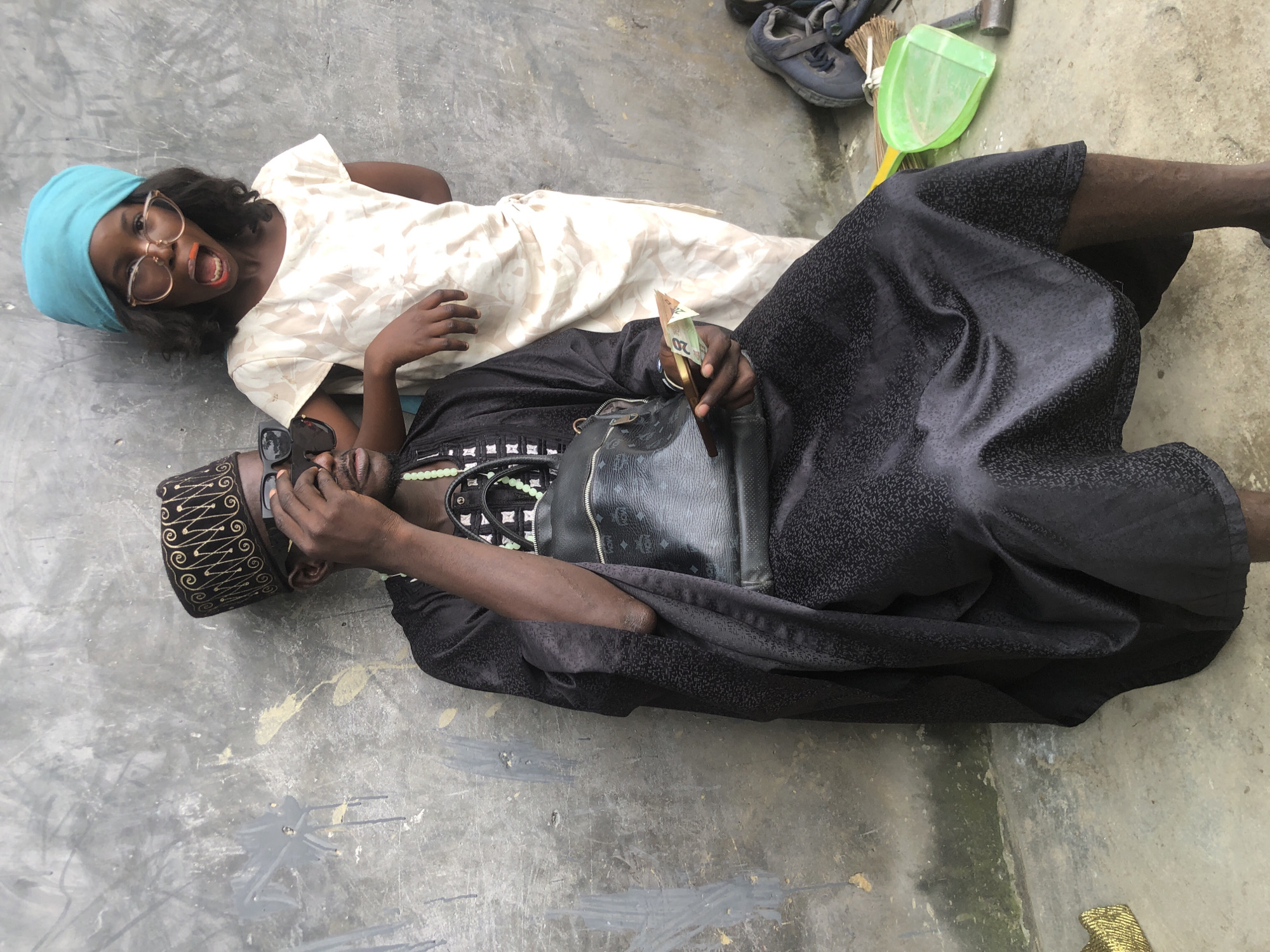 In Ado, Nigeria, a man in traditional black attire and a patterned cap sits against a concrete wall. He wears sunglasses and holds a wad of cash in his right hand. A woman or girl, adorned with a blue headscarf and large glasses, stands behind him, leaning on his shoulder with her left arm. She has bright orange lipstick and her mouth is open in a surprised expression. A black handbag hangs from the man's shoulder. In the background, a green dustpan and broom are visible.