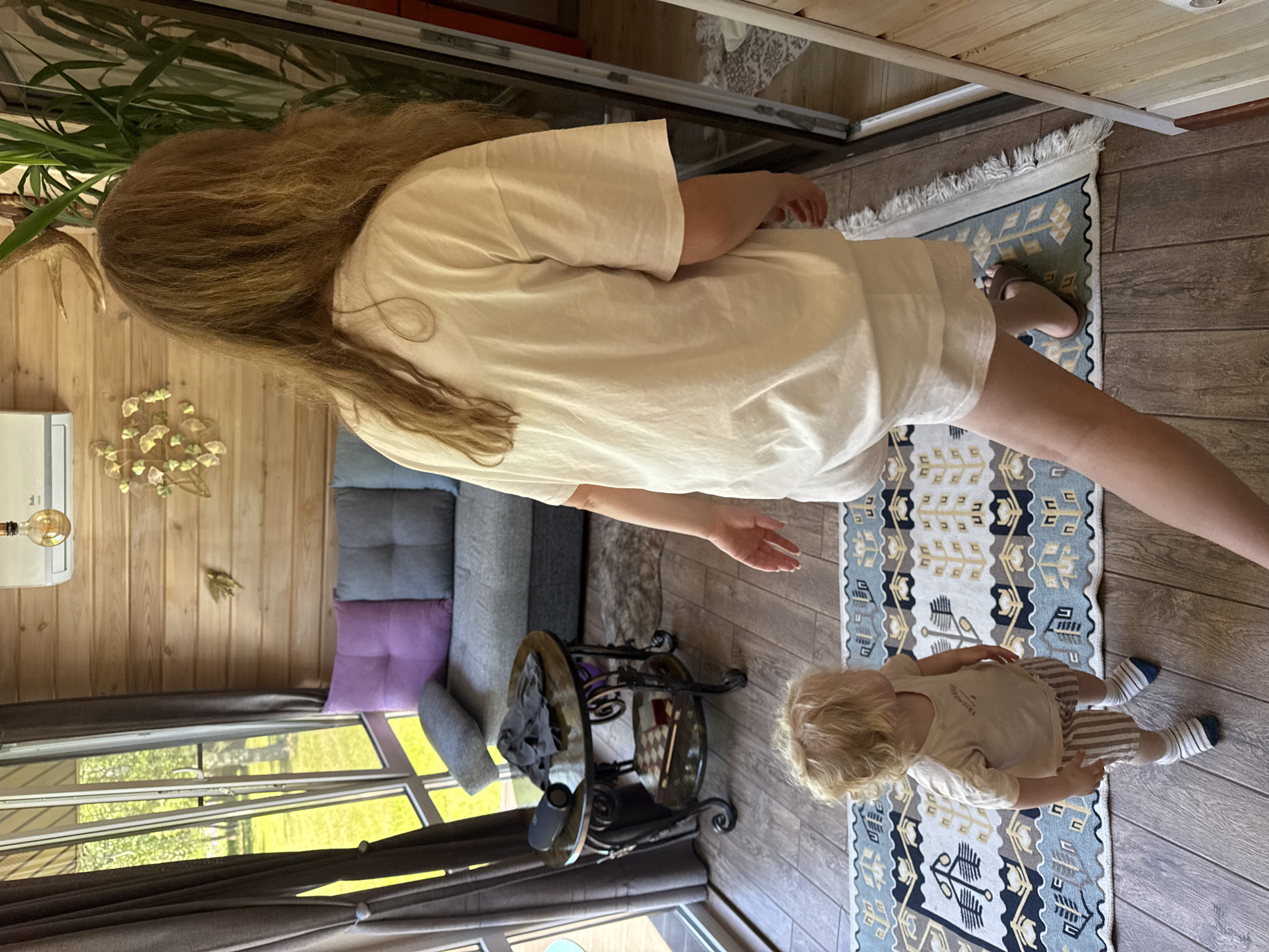 This image captures an indoor scene, specified as being in Копнино, Russia, featuring an adult female and a young child within a brightly lit room. The adult female, positioned in the foreground with her back to the camera, has long, light brown, wavy hair and wears a loose-fitting, light-colored short-sleeved t-shirt and matching shorts, with brown slide sandals. She appears to be moving towards a tall, reflective surface, possibly a mirror or glass door, on the right side of the frame. To her left, a blonde-haired toddler, also seen from a slightly elevated rear angle, stands on a decorative rug, looking towards the left. The child wears a light-colored short-sleeved t-shirt, brown and white striped shorts, and striped white and blue socks.
The room's rear wall is constructed from light-toned horizontal wooden planks. A white wall-mounted air conditioning unit is affixed to this wall, above which a decorative light bulb hangs. Below the air conditioner and to the left, two metallic wall decorations, one resembling abstract foliage and another a single leaf, adorn the wooden surface. A grey fabric sofa, featuring square tufting and adorned with one purple and one grey throw pillow, is situated against the back wall. Large, multi-pane windows on the left side of the room offer a clear view of an expansive, verdant outdoor area with trees and bright green grass. Dark grey curtains, held back by metallic ties, frame these windows.
The floor is covered with light brown wood-look planking, over which a rectangular rug with a distinctive blue, white, and brown geometric pattern, including stylized plant motifs and fringed edges, is laid in the foreground. A dark, round, two-tiered side table with ornate metal legs is positioned near the windows. On its top surface, a black portable speaker rests beside a folded dark grey garment. A wooden box, possibly a game set, occupies the table's lower shelf. A tall green plant is partially visible behind the adult female, situated near the reflective surface on the right. The room is well-illuminated by natural light entering through the large windows.