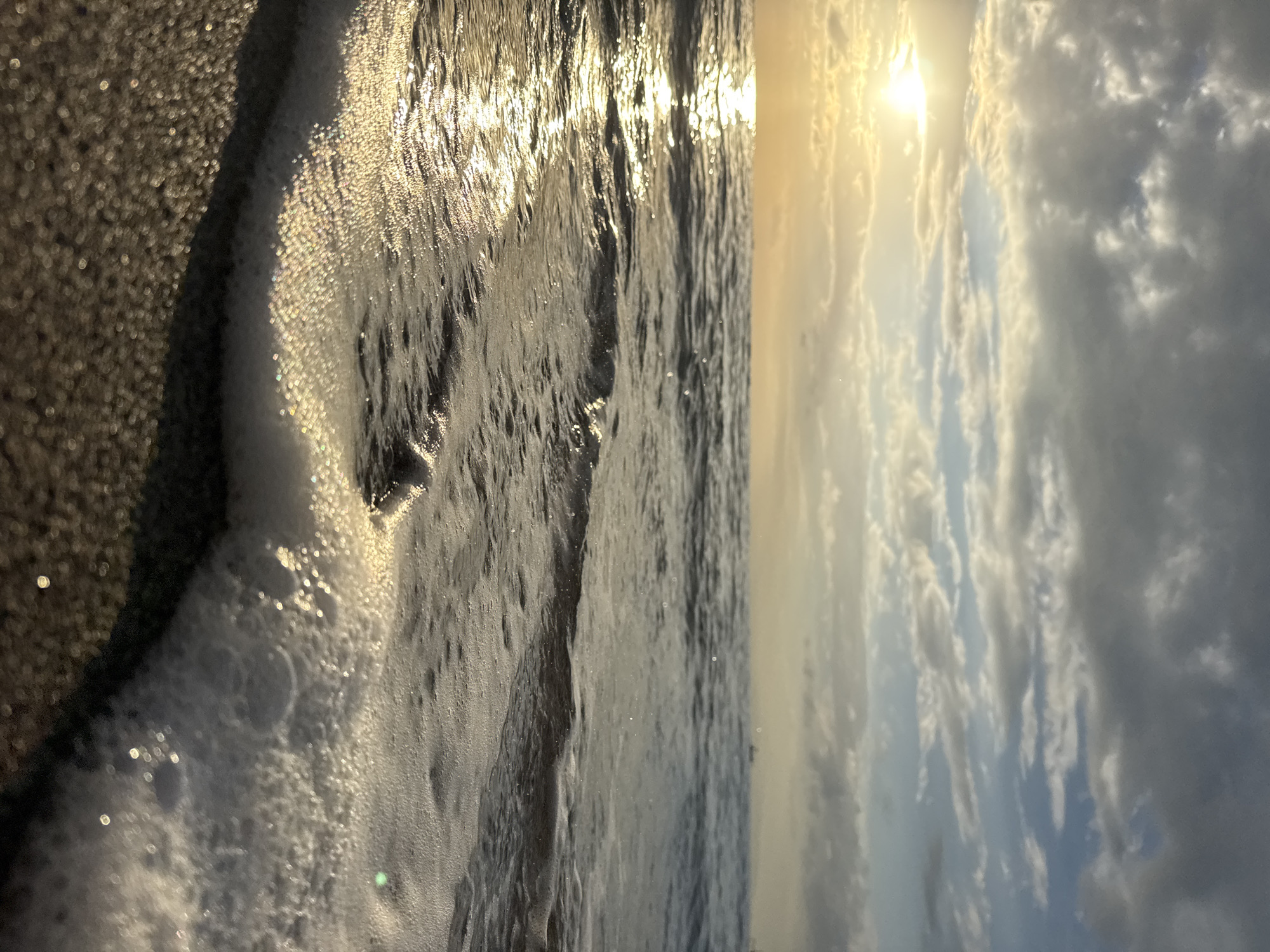This is a close-up, eye-level shot of waves gently lapping at a sandy shore during what appears to be sunrise or sunset. The sun is low on the horizon, casting a warm, golden light that reflects intensely on the water's surface. The sky is partly cloudy with a mix of grey and white clouds, interspersed with patches of blue. The foreground is dominated by wet sand and the foamy edge of a breaking wave, with small pebbles visible in the lower left. The ocean stretches out to the horizon, where distant, faint shapes that might be boats or structures can be observed. The scene conveys a sense of tranquility and natural beauty. The image does not contain any information that would suggest it is located in Abang, Indonesia.