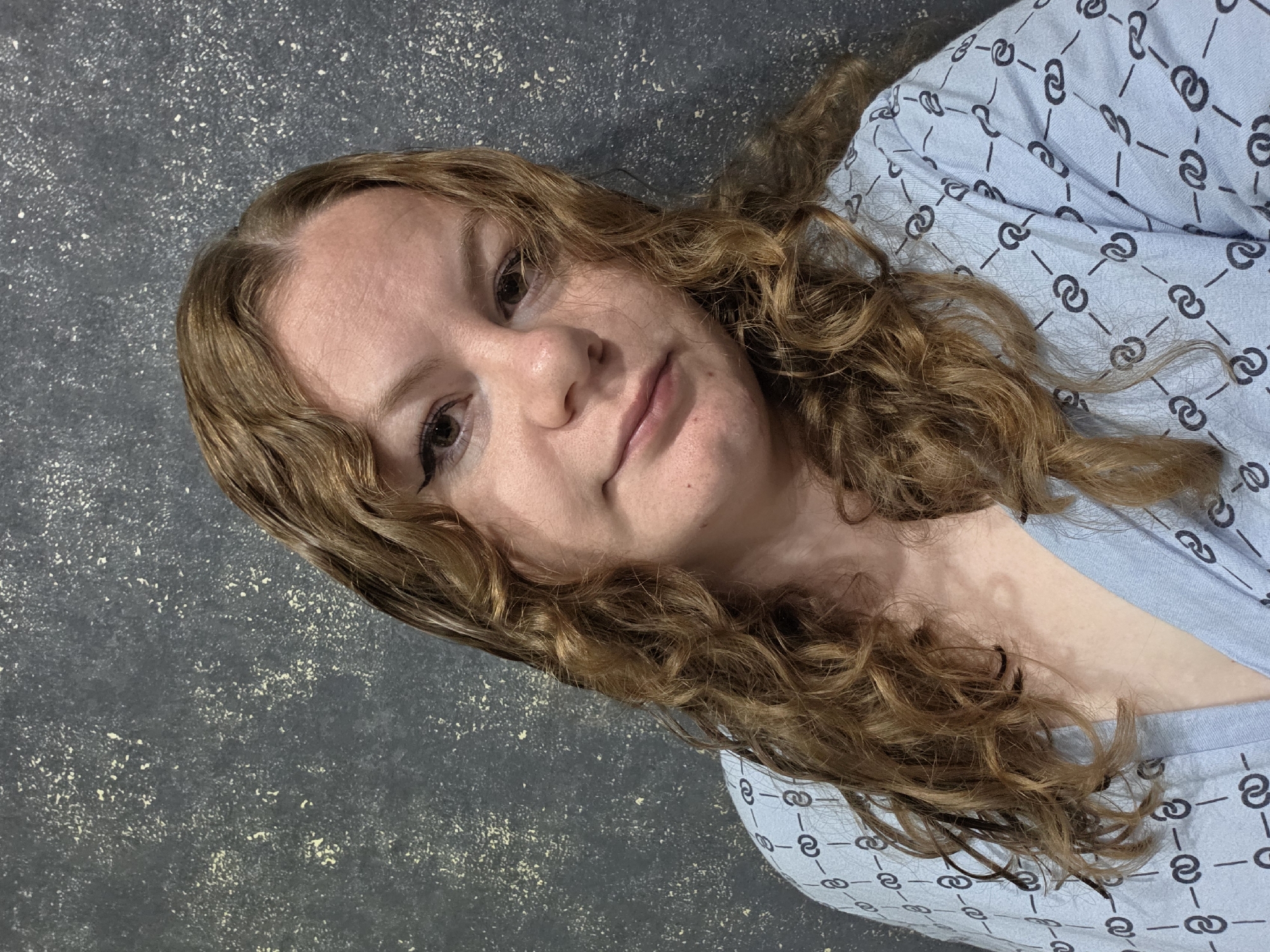 A young Caucasian woman with light brown curly hair and fair skin is pictured in a close-up, waist-up shot. She is wearing a light blue garment with a repeating pattern of black interconnected circles and lines. Her head is tilted slightly to her left, and she is looking directly at the camera with brown eyes, her lips slightly parted. She has eyeliner and what appears to be light makeup on. Her hair is loose and falls around her shoulders. The background is a dark, textured wall with speckled lighter-colored splotches. The lighting is frontal and slightly to her right. The image appears to have been taken indoors, with no obvious windows or external light sources. No specific city or location details are discernible from the image itself.