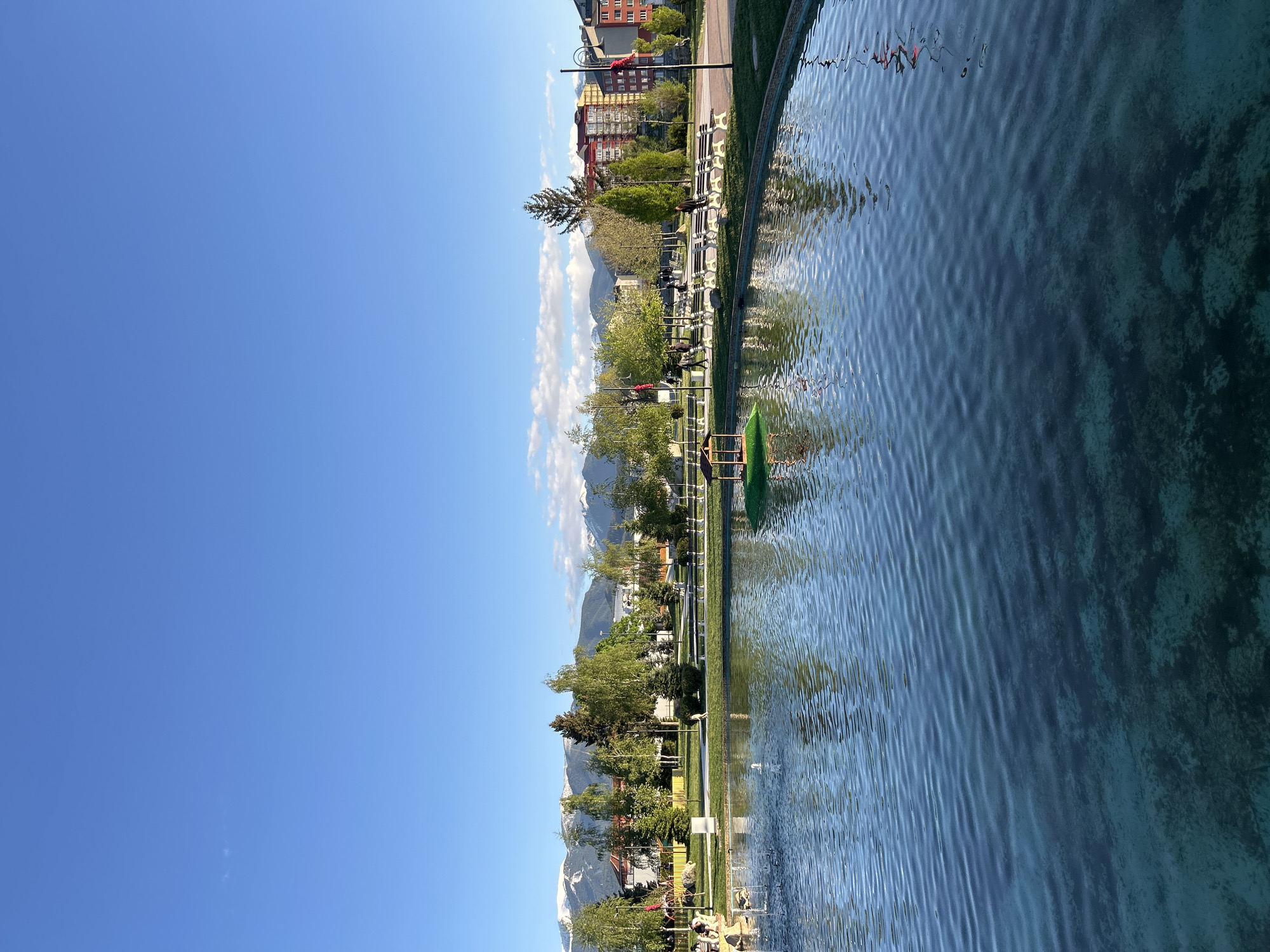 The image depicts a water scene in an urban park setting. A body of water, possibly a pond or lake, dominates the foreground. The water surface is rippled, reflecting the sky and surrounding scenery. Along the water's edge, there is a grassy area with trees, benches, and walking paths. In the background, buildings and mountains are visible. The sky is clear blue with some scattered clouds. The overall impression is one of a peaceful and picturesque urban park landscape.