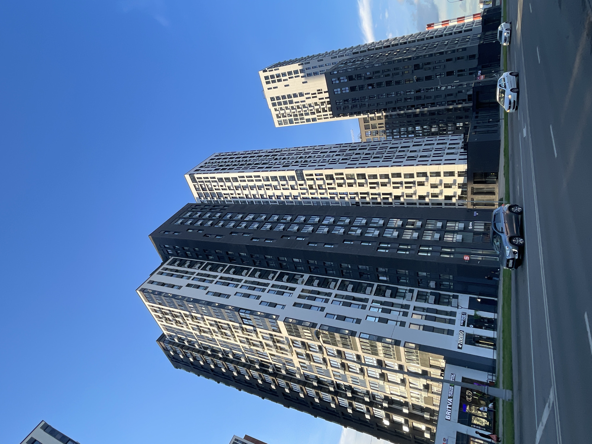 The image shows a wide shot of a modern residential complex in Moscow, Russia, under a clear blue sky. The buildings are tall, multi-story structures with striking architectural designs, featuring a combination of dark and white facades with a grid-like pattern of windows. Sunlight casts shadows on the buildings, suggesting it is late afternoon or early evening.
In the foreground, a road runs alongside the buildings, with several cars visible. One dark blue car is parked on the right side of the road, and two white cars are driving on the left side, moving away from the viewer. The road has lane markings, indicating a street or highway.
The lower sections of the buildings appear to have commercial spaces with visible signage. Text in Russian can be seen on some of the storefronts, including "ПИВО" (beer) and "BRITVA" (razor), implying a mix of businesses catering to residents.
The scene conveys a sense of urban development and modern living in a bustling city. The overall mood is calm and orderly, with the clear sky and bright sunlight contributing to a pleasant atmosphere. The perspective is from a low angle, looking up at the imposing architecture, emphasizing the scale of the buildings.