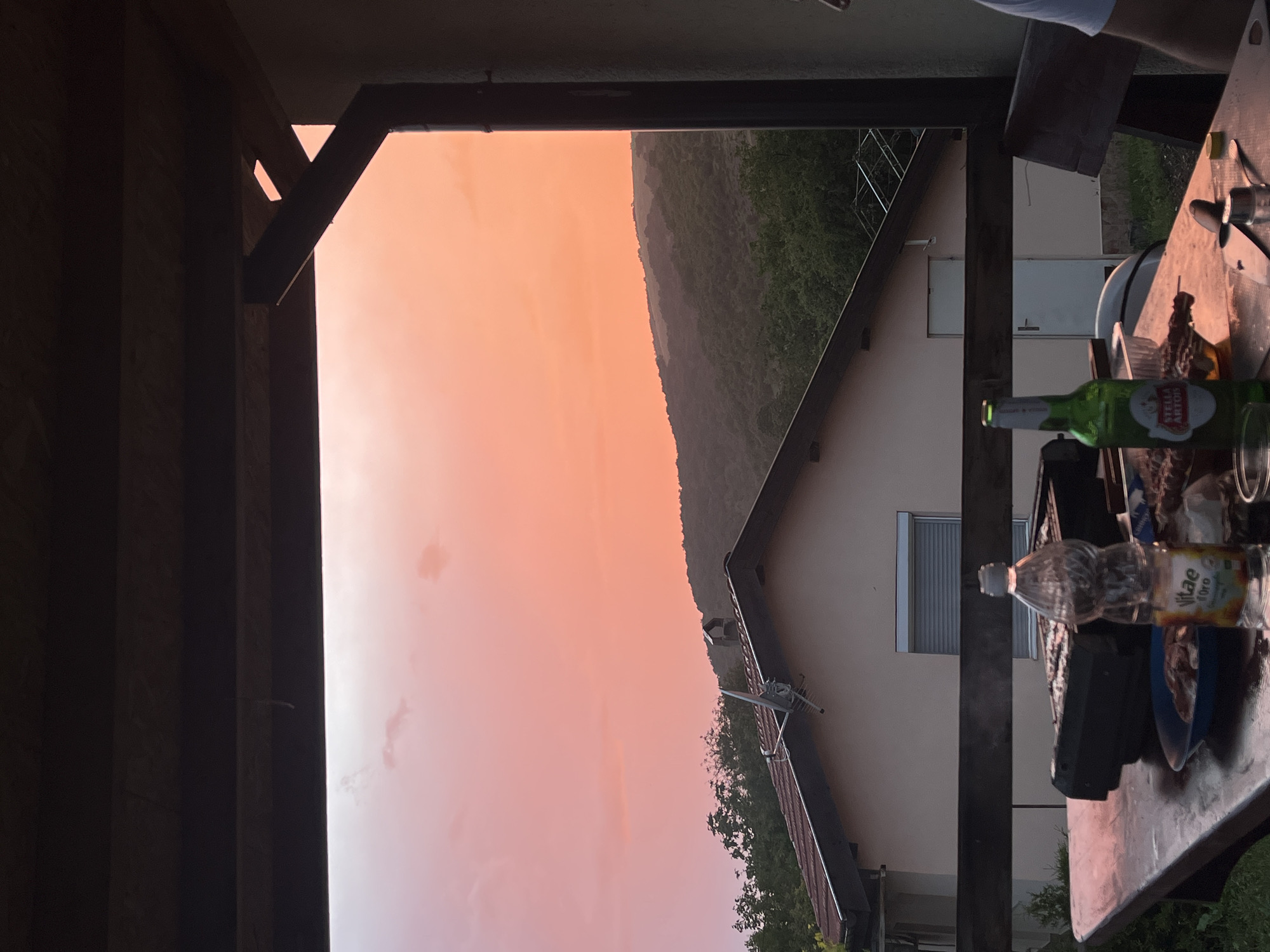 The image, oriented sideways, captures an outdoor scene at sunset or sunrise from under a dark wooden structure, possibly a porch or balcony, in Kosten, Bulgaria. The sun's low angle casts a warm, vibrant orange and pink glow across a vast, clear sky, dominating the left side of the frame.
In the foreground, on the right side of the rotated image, is a wooden table laden with items. A green glass bottle prominently displays "Stella Artois" on its label, and a clear plastic bottle appears to be "Vitae Oro" water. Other indistinct objects, possibly food or utensils, are scattered on the table, suggesting an outdoor meal or gathering. Part of a person's arm, wearing a white short-sleeved shirt, is visible at the very top right, indicating someone is seated or standing nearby. Dark wooden beams and posts of the overhead structure frame the scene on the top and bottom.
Beyond the immediate foreground, in the mid-ground, a light-colored house with a dark-tiled roof and a window with horizontal blinds is visible. An antenna or aerial protrudes from the roof. Lush green foliage and trees are scattered around the house and extend towards the background.
The background, on the left side of the image, features a large, dark green, tree-covered hill or mountain range silhouetted against the bright sky. The overall impression is one of a relaxed evening or morning gathering, enjoying drinks and possibly food, with a scenic view as the sky transitions through vivid colors.