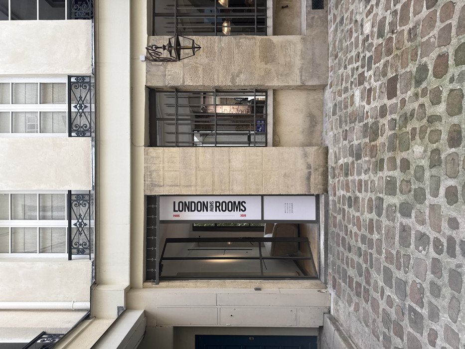 The image depicts an interior courtyard in Paris, France, paved with irregularly shaped cobblestones. The focal point is the ground-floor entrance of a building, featuring a modern, dark-framed glass double door, partially ajar, revealing a well-lit interior with a cream-colored armchair. To the right of the entrance, a vertical white banner is affixed to the stone wall, displaying "LONDON ROOMS" prominently in black uppercase letters, with "PARIS" and "MOHS" above, and "BRITISH FASHION COUNCIL" and "2025" below.
The building's facade is constructed from rough-hewn, light-colored stone blocks on the ground level, transitioning to a smooth, cream-colored stucco on the upper floor. Above the ground-floor entrance and windows are three rectangular windows on the upper floor, each fitted with white frames, sheer curtains, and decorative black wrought-iron balconies. Two ground-floor windows to the right of the entrance are secured with vertical black metal bars; an ornate, dark-colored wall-mounted lantern with glass panes hangs between these windows. To the far left, a dark blue single door is set into a recess, adjacent to a plain beige wall. The overall scene suggests a blend of historical Parisian architecture with contemporary design elements, likely indicating a showroom or event space.
