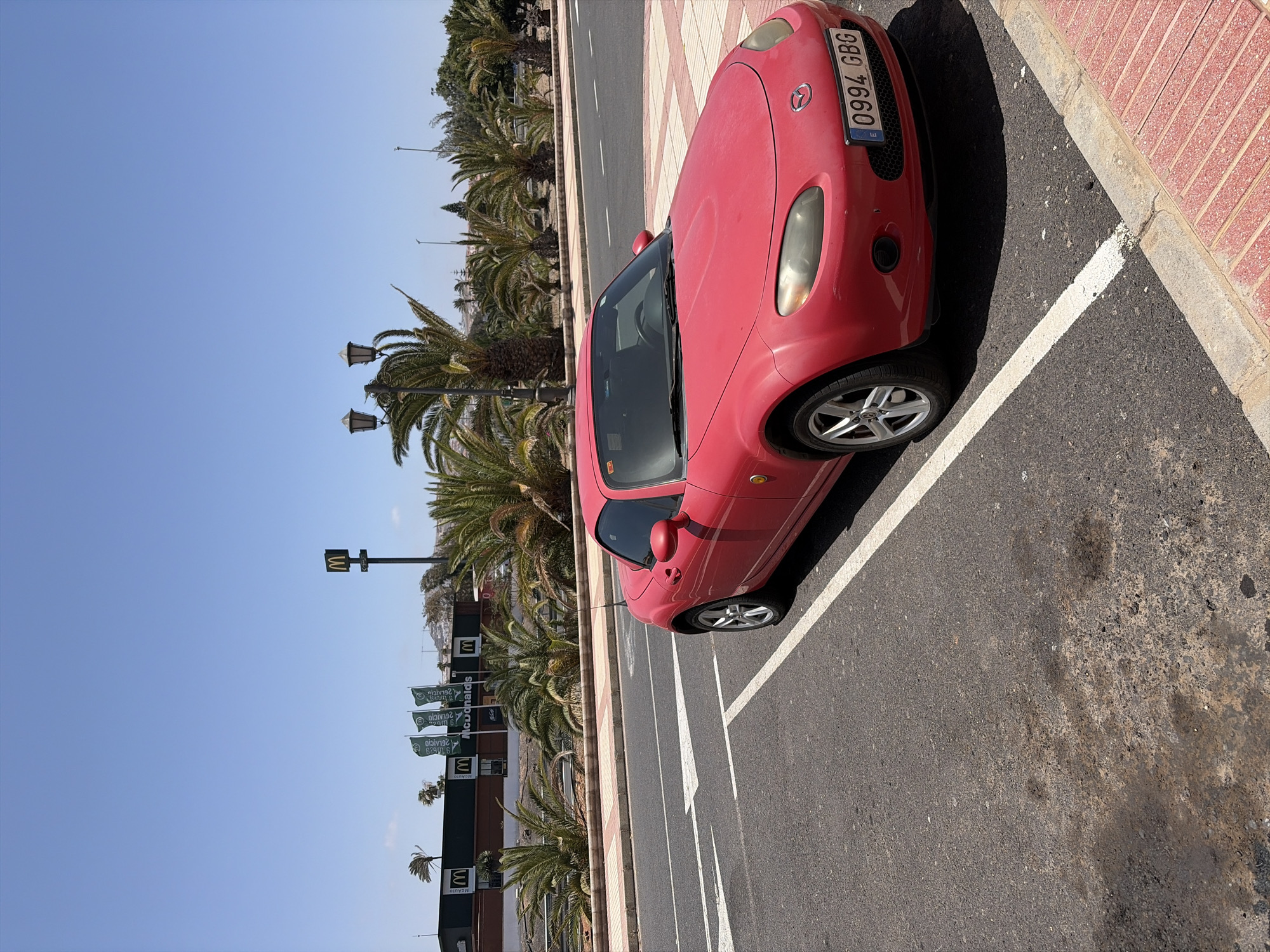 A red Mazda convertible is parked in a parking space in front of a McDonald's restaurant. The McDonald's has a green facade and several green flags with the McDonald's logo. There are palm trees lining the street in front of the restaurant. The sky is clear and blue. The license plate on the car reads "0994 GBG".