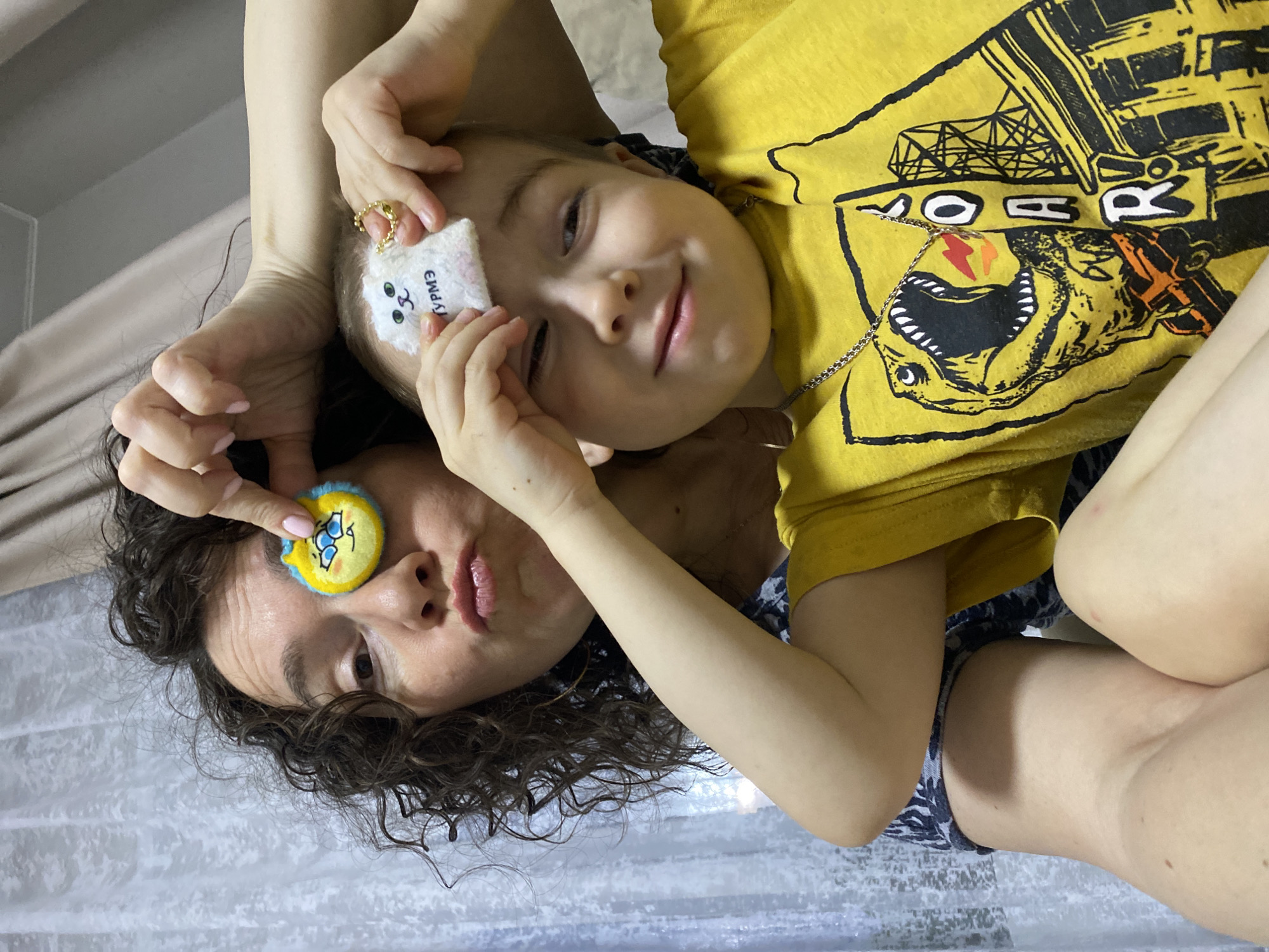 A woman and a child are posing for a picture. The woman has curly brown hair and is holding a yellow and blue object to her eye. The child is wearing a yellow shirt with a dinosaur on it and holding a white object with a face to their eye. The photo was taken indoors. The location is unknown, and there is no evidence to suggest it was taken in Noviy, Russia.