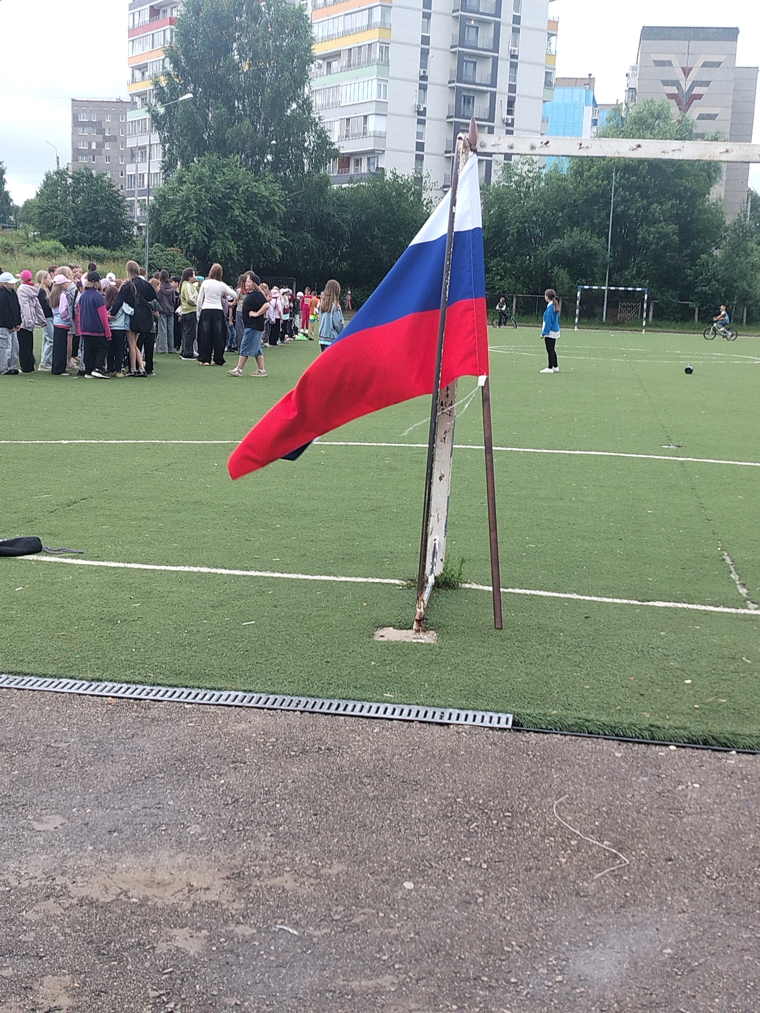 The image depicts an outdoor sports field in Chaikovsky, Russia, likely a schoolyard or public park given the presence of children and residential buildings in the background. A Russian flag is prominently displayed in the foreground, mounted on a pole. Several groups of children are visible, some standing in rows, possibly waiting for an event or activity. Two individuals are seen riding bicycles in the background on the field. The environment includes a green artificial turf surface typical of sports fields, with white lines marking boundaries. Adjacent to the field is a paved area with a drainage grate. The background features multi-story apartment buildings, characteristic of urban or suburban Russian architecture, and mature trees. The sky is overcast, suggesting a cloudy day.