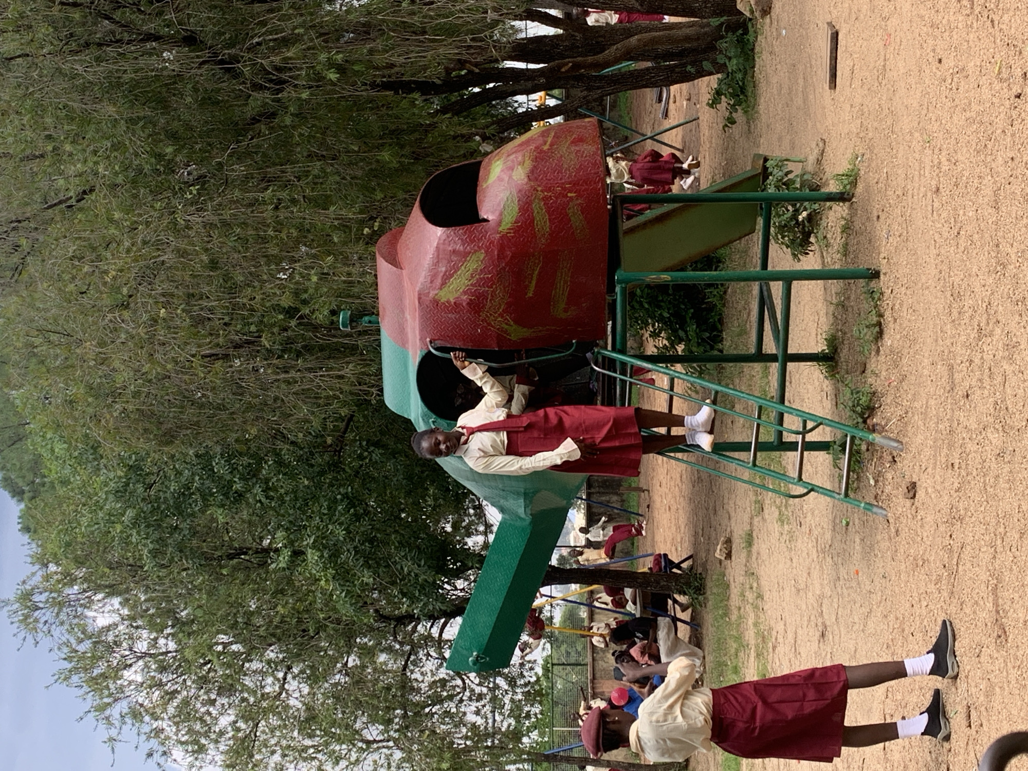 In Jos, Nigeria, a group of schoolchildren are gathered around a playground structure designed to resemble a helicopter. A young girl in a school uniform stands on a ladder leading up to the helicopter structure, looking towards the right. Her uniform consists of a cream-colored shirt, a red tie, and a red skirt. Another student, also in uniform, stands in the foreground, facing away from the camera, holding a phone and seemingly taking a picture of the helicopter. This student wears a cream-colored shirt, a red skirt, and a red cap. The playground is covered in sand, and there are trees and other children in the background. One child is on a swing set in the mid-ground, while others are seated at a picnic table. The helicopter structure itself is painted red with green accents and appears to be a piece of play equipment.