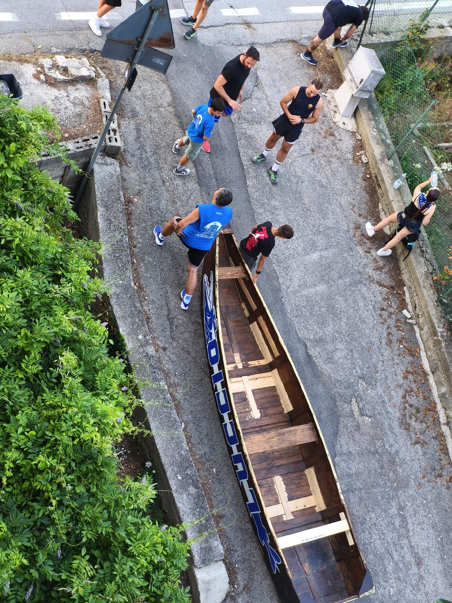 The aerial photograph, taken in Passignano sul Trasimeno, Italy, depicts a group of people standing on a paved surface next to a wooded area and a body of water. The scene appears to be a street or pathway, possibly near a park or waterfront. In the foreground, a long, narrow wooden boat is positioned on the paved surface, its interior visible. The boat has blue lettering along its side.
Several individuals are present. A person in a blue sleeveless shirt and black shorts, possibly a runner, is standing near the boat. To their left, a younger person in a blue t-shirt and shorts is visible, also on the paved surface. Further up the pathway, a group of individuals in athletic wear are gathered. One person in a black t-shirt and shorts is bending over. Another person in a black tank top and shorts is holding a water bottle. Further back, two individuals in dark athletic clothing are standing. One appears to be interacting with a signpost.
The environment includes lush green foliage on the left side of the image, suggesting a natural or park-like setting. A concrete barrier runs alongside the paved area, and a wire fence can be seen further in the background. Scattered leaves are present on the ground. The lighting suggests it is daytime, with no obvious shadows indicating a specific time of day. The overall impression is a casual outdoor gathering, possibly related to a sporting event or recreational activity.
