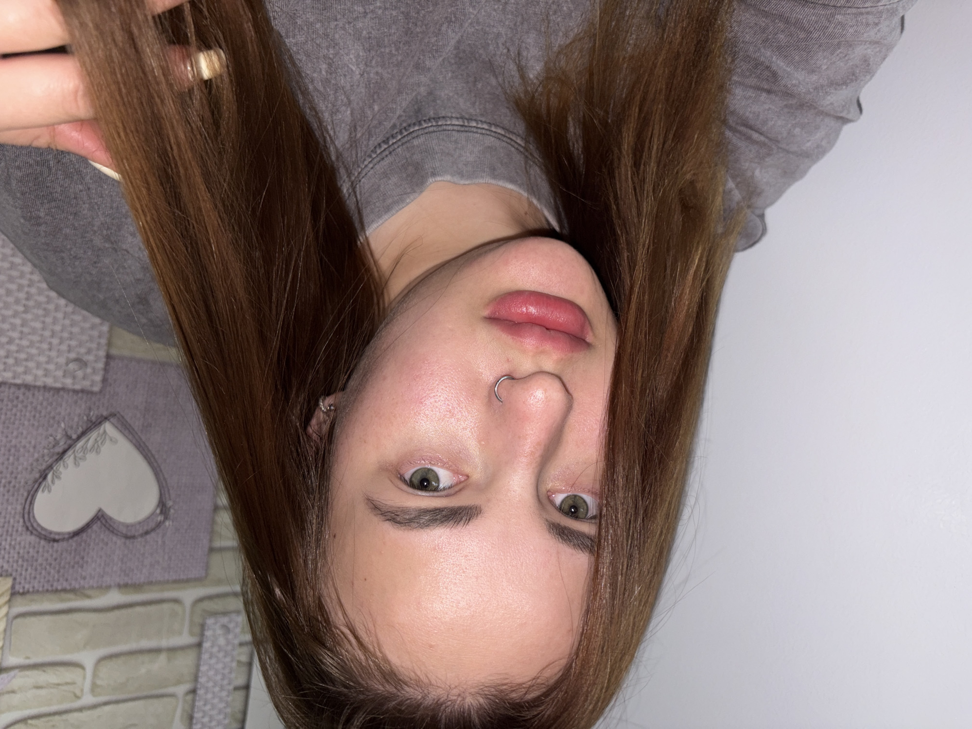A photograph shows a female individual oriented upside down, likely taken indoors. The subject has light skin, brown hair, and green eyes. A piercing is visible in her left nostril. She is wearing a gray sweater. The background is divided: one side is a plain white wall, and the other side features a wall with a heart-shaped decoration and a brick-like pattern. The location and city are not discernible from the image.