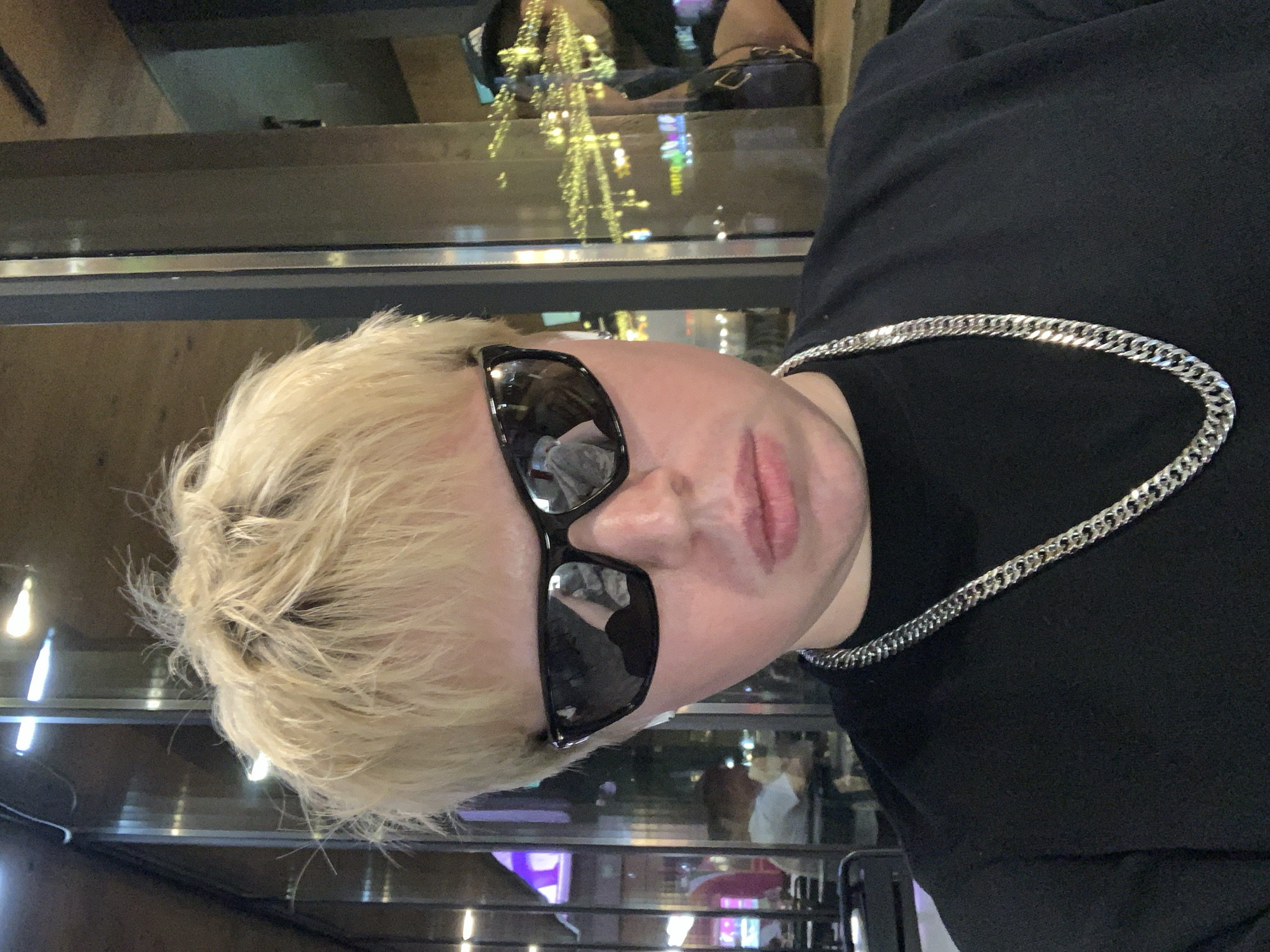 A medium, vertically-oriented shot depicts a person with blonde hair wearing black sunglasses and a silver chain necklace. They are wearing a black shirt. The background is a blurred view of a cityscape reflecting off a window. The location is in Almaty, Kazakhstan, suggested by the presence of buildings and reflections within the cityscape. The overall impression is of an indoor scene, possibly a restaurant or cafe, with the person posing for the photo against the window.