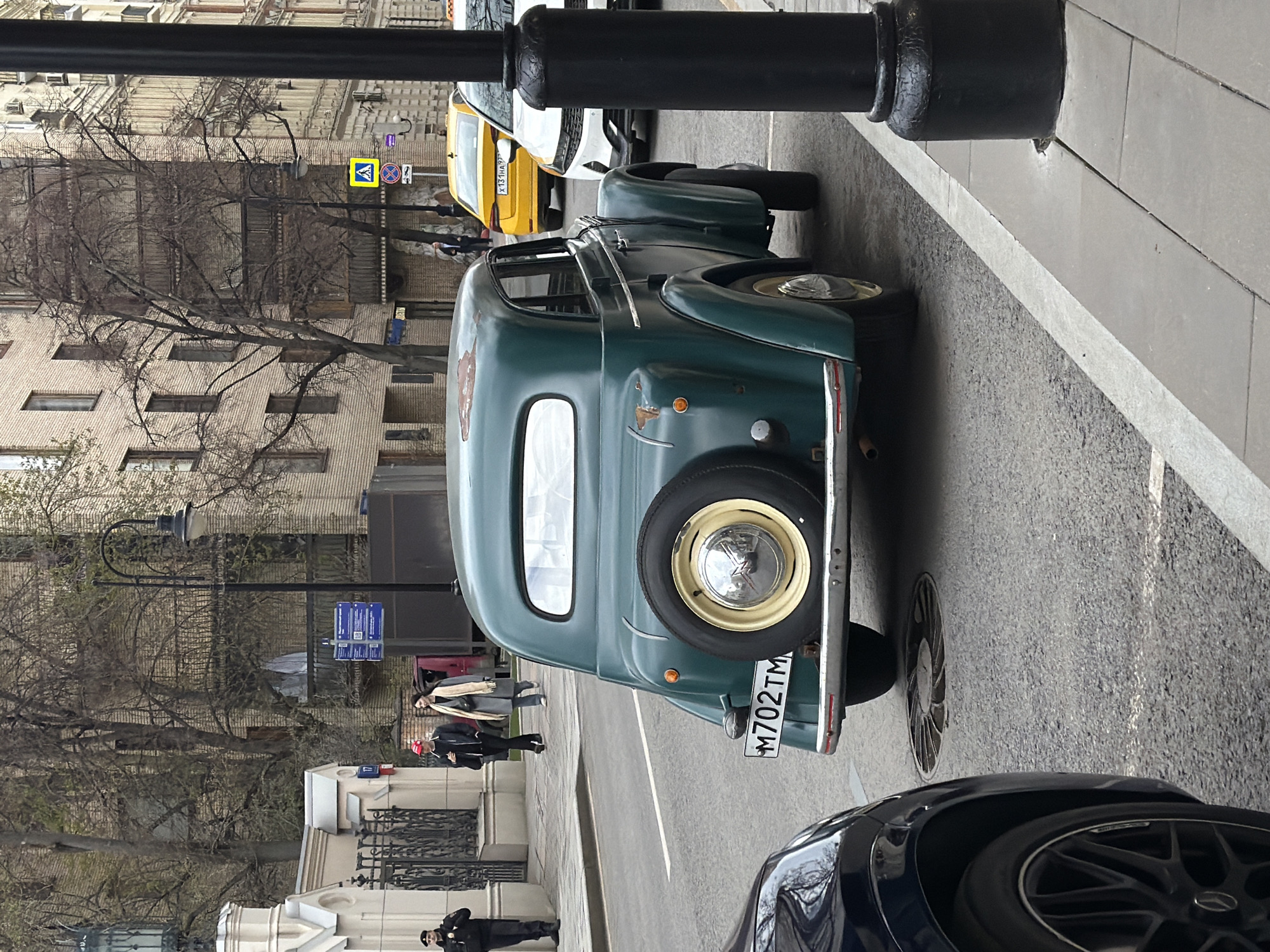 An outdoor street scene in Moscow, Russia, is depicted during what appears to be an overcast daytime. The image focuses on a vintage dark green car, possibly a GAZ-M20 Pobeda, parked parallel to a sidewalk on the right side of the street. The car is seen from the rear, revealing a spare tire mounted on its trunk and a license plate that reads "M702TM". The vehicle shows visible signs of wear and rust.
In the immediate foreground, the front wheel and part of the hood of another dark-colored car are visible. A black lamppost or utility pole stands prominently on the right. The street is paved with asphalt, and a circular grate is visible on the road surface near the vintage car.
Beyond the parked car, a sidewalk runs alongside buildings. Several pedestrians are visible on the sidewalk: a person in a black jacket and red hat, another in a long grey coat and scarf, and a person in uniform (possibly a security guard or police officer) standing near a building with classical architectural details. Bare trees with intricate branches line the street, suggesting late winter or early spring. In the background, other cars, including a yellow taxi and a white car, are visible. Street signs, including a yellow pedestrian crossing sign and a blue informational sign with QR codes, are also present but largely illegible.