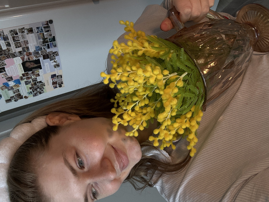 The image depicts a young adult female holding a clear, faceted glass pitcher containing a bouquet of yellow, fluffy flowers, consistent with mimosa. The woman, who appears to be taking a self-portrait, has fair skin, light-colored eyes, and light brown hair partially pulled back by a pale, textured headband. She wears a light-colored, vertically striped or ribbed collared shirt and exhibits a slight, downward-gazing smile. Her right hand is visible, gripping the handle of the pitcher.
The background reveals a light-colored refrigerator, prominently adorned with a collage of numerous small photographs and paper notes. Several notes display Cyrillic text, including "ЗДОРОВЬЕ" (Health), "СЕМЬЯ" (Family), "КАРЬЕРА" (Career), "ПУТЕШЕСТВИЯ" (Travels), "МЕЧТЫ" (Dreams), and "ДЕТИ, ТВОРЧЕСТВО" (Children, Creativity). A larger, central photograph of a woman's face is affixed to the refrigerator.
The scene takes place indoors, within a residential environment, likely a kitchen or a room with a refrigerator. The specific location within the city of Nikol'skii, Russia, cannot be determined from the visual evidence; however, the Cyrillic text confirms a Russian-speaking context. Lighting is soft and even, illuminating the subject and the immediate background.