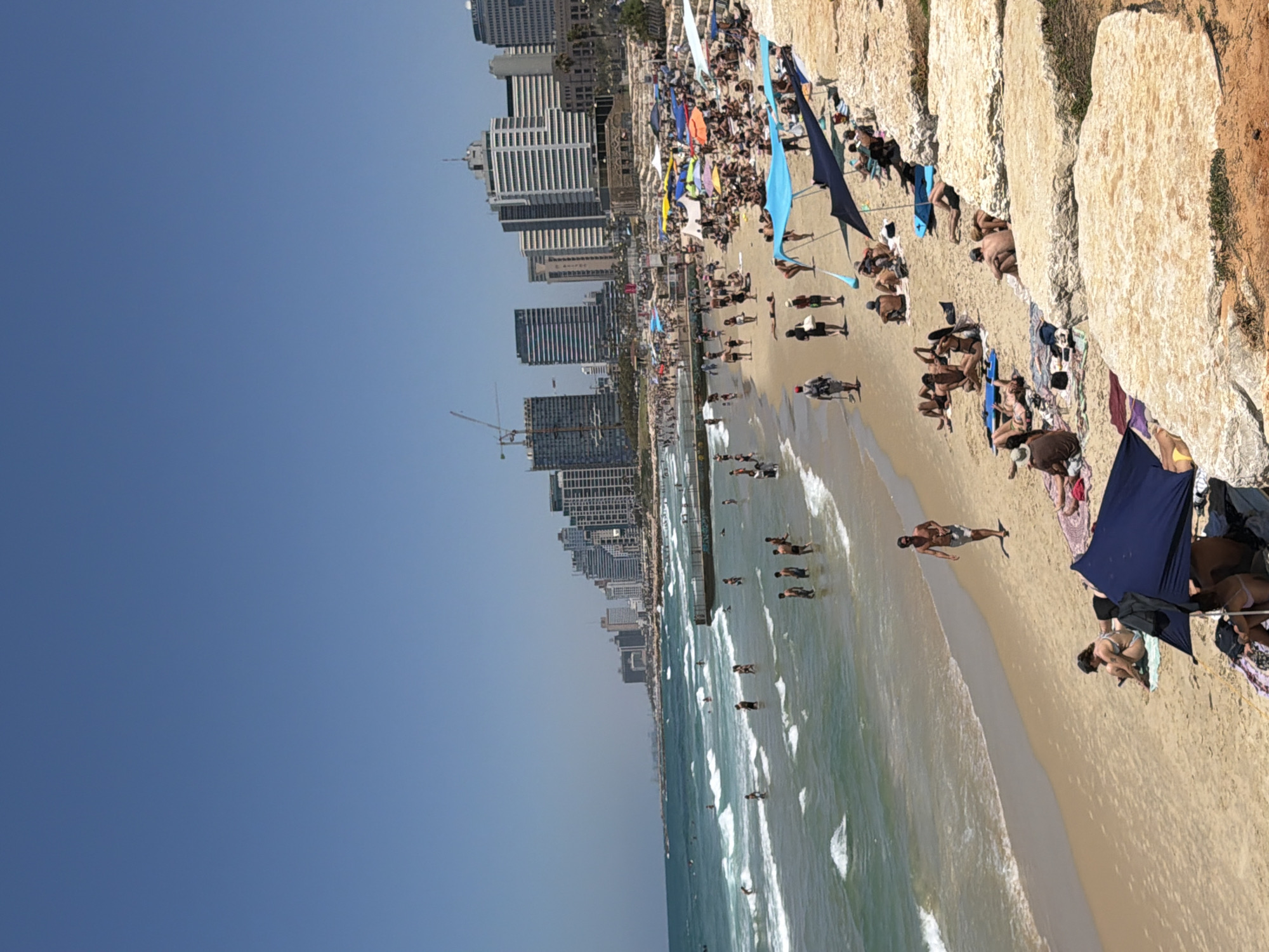 The image shows a beach scene in Tel Aviv, Israel. The beach is crowded with people sunbathing, swimming, and walking along the shoreline. There are several colorful umbrellas providing shade. In the background, a line of modern buildings forms the Tel Aviv skyline. The sky is clear and blue. A rocky cliff runs along a portion of the beach.
