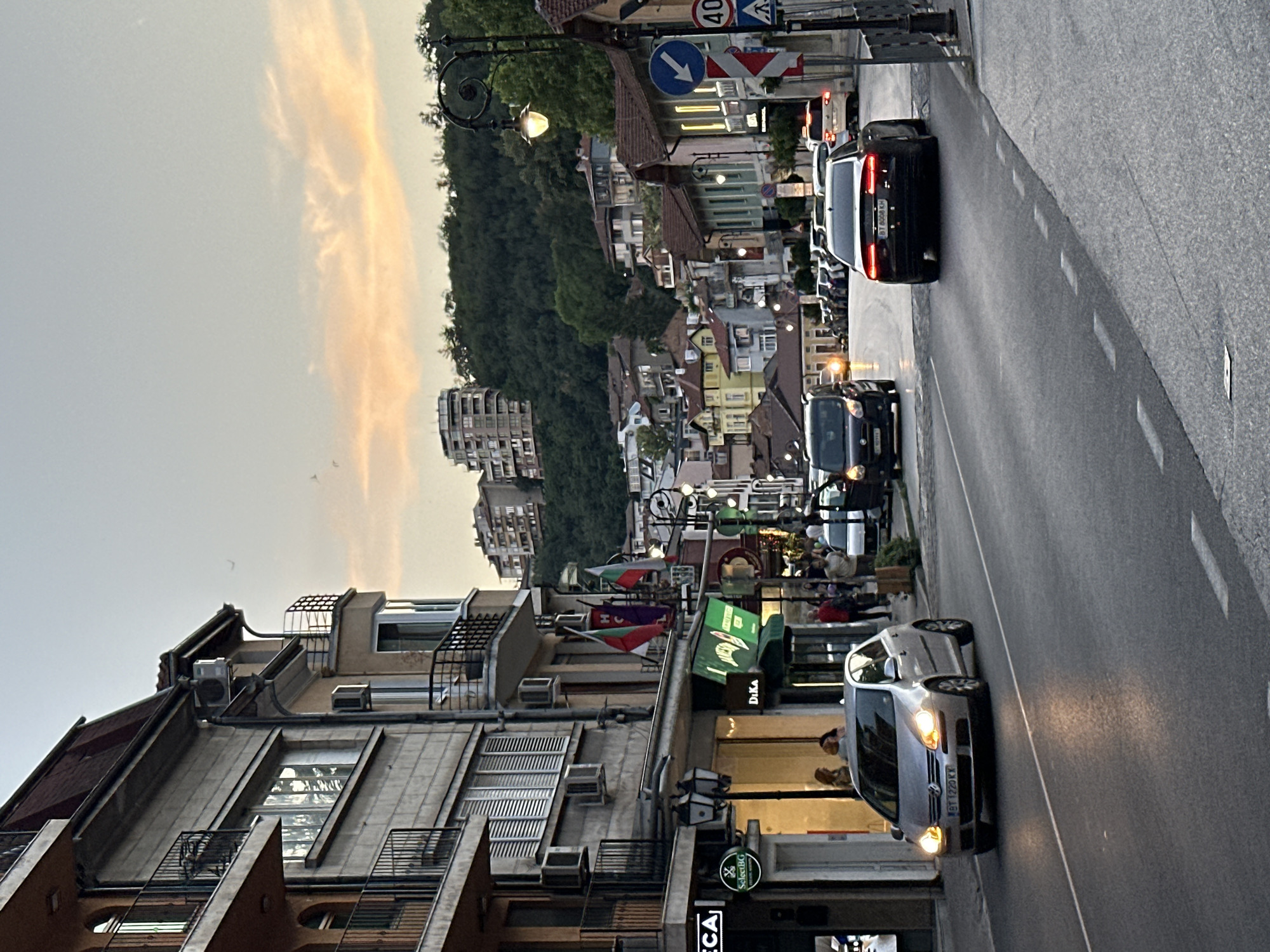 The image depicts a street scene in Veliko Tarnovo, Bulgaria, during what appears to be dusk or early evening. The perspective is from the middle of a paved road looking uphill towards a densely built area with numerous buildings of varying architectural styles, predominantly in shades of beige, yellow, and light green. Many of the buildings are multi-story, with balconies and tiled roofs. Lush green trees and vegetation cover the hillside behind the buildings.
Several cars are visible on the road, including a silver Volkswagen Golf in the foreground on the left, a dark-colored van in the middle, and a dark sedan with illuminated taillights further up the road. White lines delineate the traffic lanes.
On the left side of the street, a commercial building features shops with illuminated storefronts and awnings. Flags, including the Bulgarian flag, are displayed outside one of the establishments. People are seen walking on the sidewalk in front of the shops.
On the right side of the street, a lamppost with a decorative scrolled iron base and an illuminated lamp stands prominently. Road signs are also visible, including a blue circular sign with a white arrow pointing left, and a circular sign with the number "40". The sky above is a soft gradient of pale blue and peachy orange, suggesting the setting sun. The overall impression is of a lively urban environment nestled within a hilly landscape.