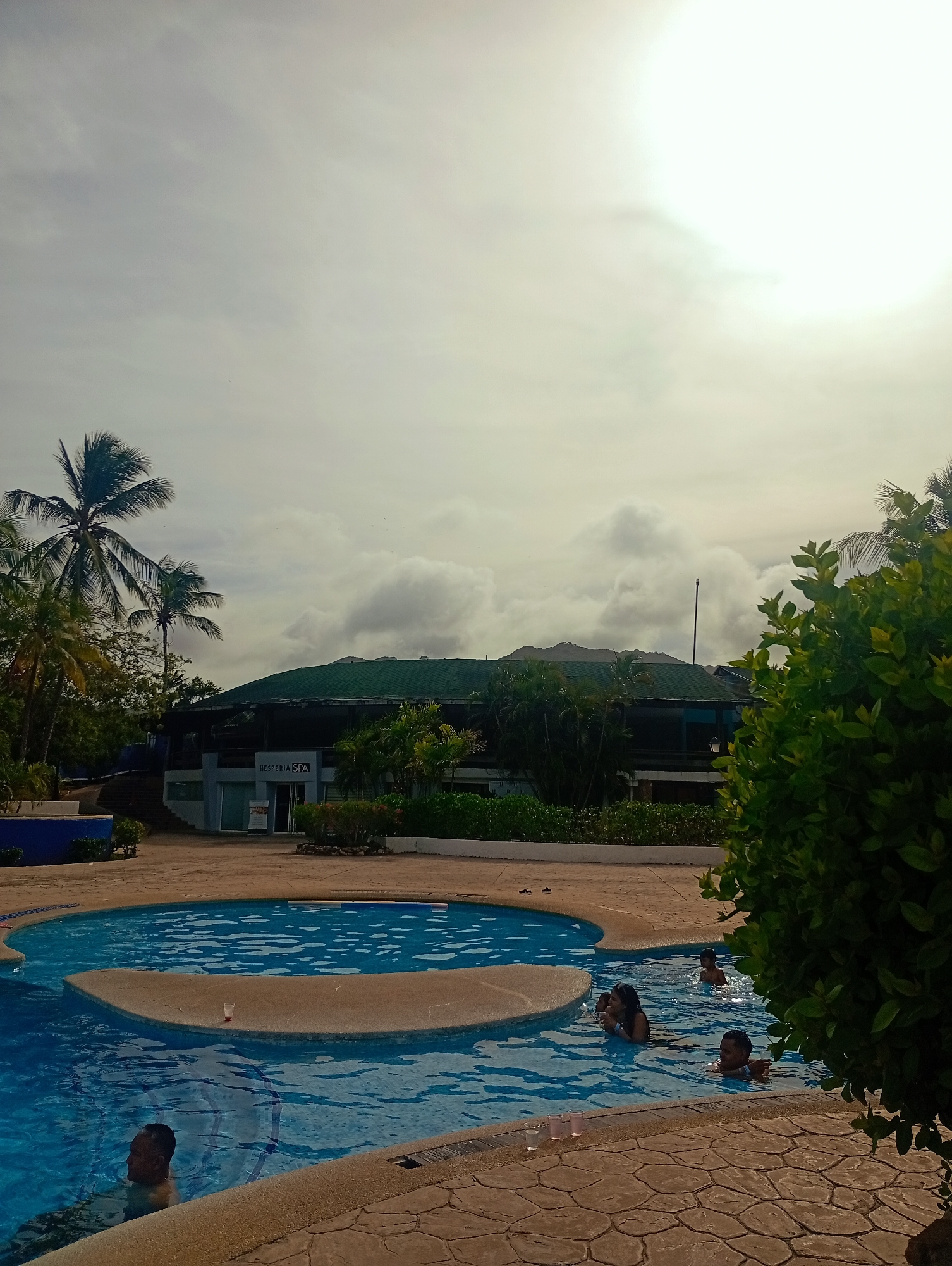 The image shows a swimming pool area in Playa El Agua, Venezuela. Several people are swimming and standing in the pool. Palm trees are visible on the left and behind a building that looks like a spa. The sky is overcast with clouds. The pool area has a paved border. Some drinking glasses are on the edge of the pool.