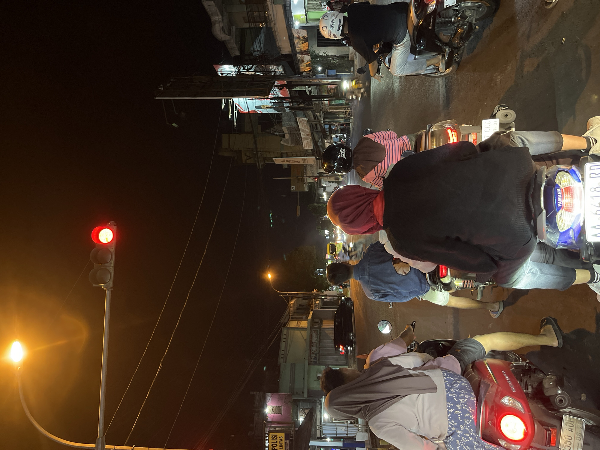 At night, a group of people on motorcycles are stopped at a traffic light in the city of Kalinyamatan, Indonesia. The traffic light is red, indicating that vehicles should stop. The street is illuminated by streetlights and the headlights of vehicles, casting long shadows on the asphalt. Buildings line the street on both sides, some with illuminated signs. The people on the motorcycles are wearing casual clothing and helmets for safety. They appear to be waiting patiently for the light to change. The atmosphere is calm and orderly, despite the darkness.