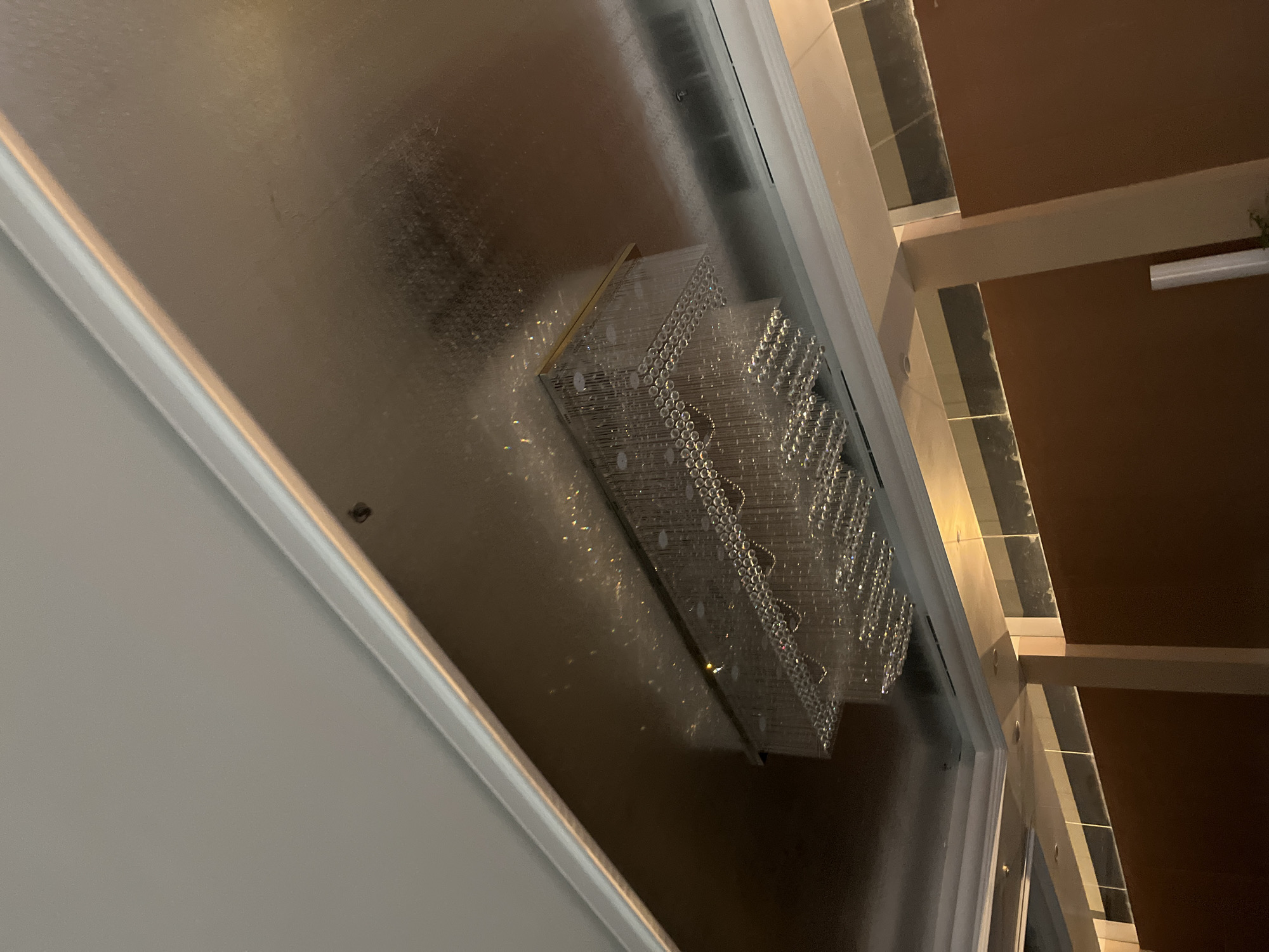 The image captures a close-up, low-angle view of a rectangular crystal chandelier hanging from a metallic ceiling. The chandelier is tiered, with multiple rows of clear crystal prisms. The ceiling is reflective and appears to have a metallic finish, casting a reflection of the chandelier. The edges of the ceiling are trimmed with white molding. Below the ceiling, there are brown walls and columns, suggesting an indoor setting. The lighting is dim, with the chandelier being the primary light source, creating numerous reflections and refractions on the ceiling. The overall impression is one of an opulent interior. The image's metadata does not specify a location in Binong, Indonesia, nor are there any people or discernible actions depicted.