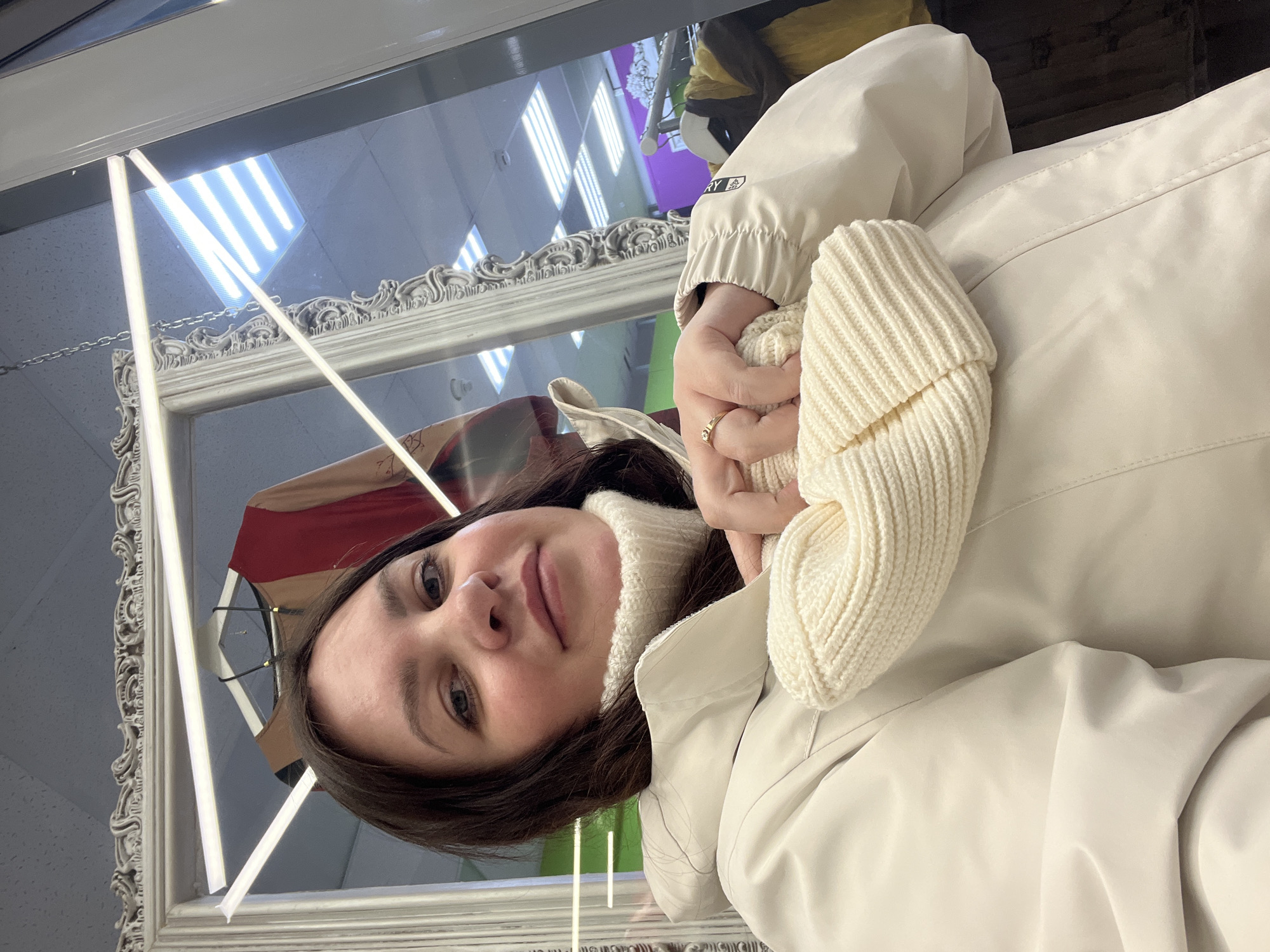 A young woman with dark hair and light eyes is captured in an indoor setting, likely a shop or boutique in Perm, Russia. The image is oriented vertically, showing a close-up of her from the chest up as she looks directly at the camera with a calm, subtle smile.
She is dressed in a light cream or off-white ribbed, high-neck knitted sweater, over which she wears a light beige or cream jacket. Her hands are clasped together at chest level, mostly enveloped by the long, ribbed cuffs of her sweater. A gold-colored ring with a small, clear stone adorns her right ring finger. On the left sleeve of her jacket, a small rectangular patch with a logo and the partial text "DISCOVERY" is visible.
The woman is positioned in front of a large, ornate mirror with a decorative, possibly carved or molded, light-colored frame. The mirror's reflection reveals elements of the surrounding commercial space: a ceiling with bright rectangular fluorescent light panels, and a clothing rack in the background holding various garments, one of which appears reddish-brown. The scene suggests she is posing for a photo, possibly a selfie, within a retail environment.