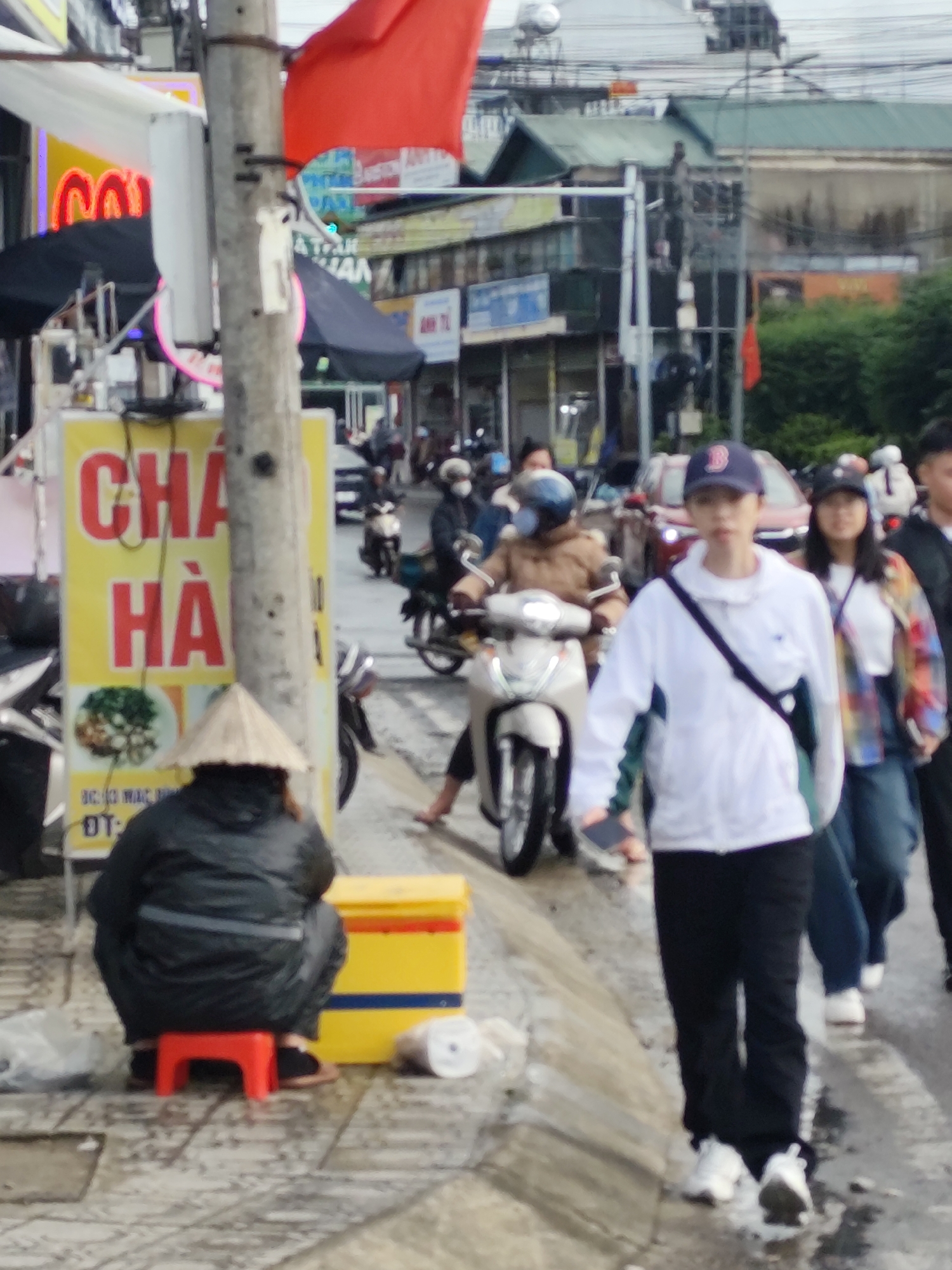 This image depicts a street scene in Da Lat, Vietnam, likely during overcast weather given the damp appearance of the pavement. In the foreground, a person wearing a conical hat and a dark jacket is seated on a small red stool. Adjacent to them is a yellow cooler with blue and orange trim.
To the left, a yellow sign with large red lettering reading "CHA HÀ" is visible, along with an image of food. A utility pole stands prominently to the left of the sign, with wires and a red flag attached to it.
In the midground, several individuals are on motorcycles. One person in a light brown jacket and helmet is riding a white scooter. Another individual in the background, wearing a white jacket and a dark baseball cap, is walking and holding a dark object, possibly a phone. A woman in a colorful plaid jacket is walking behind them.
The background shows buildings with green roofs and a complex network of electrical wires overhead. The street appears wet, suggesting recent rain. The overall impression is of a busy urban environment with pedestrian and vehicle traffic.
