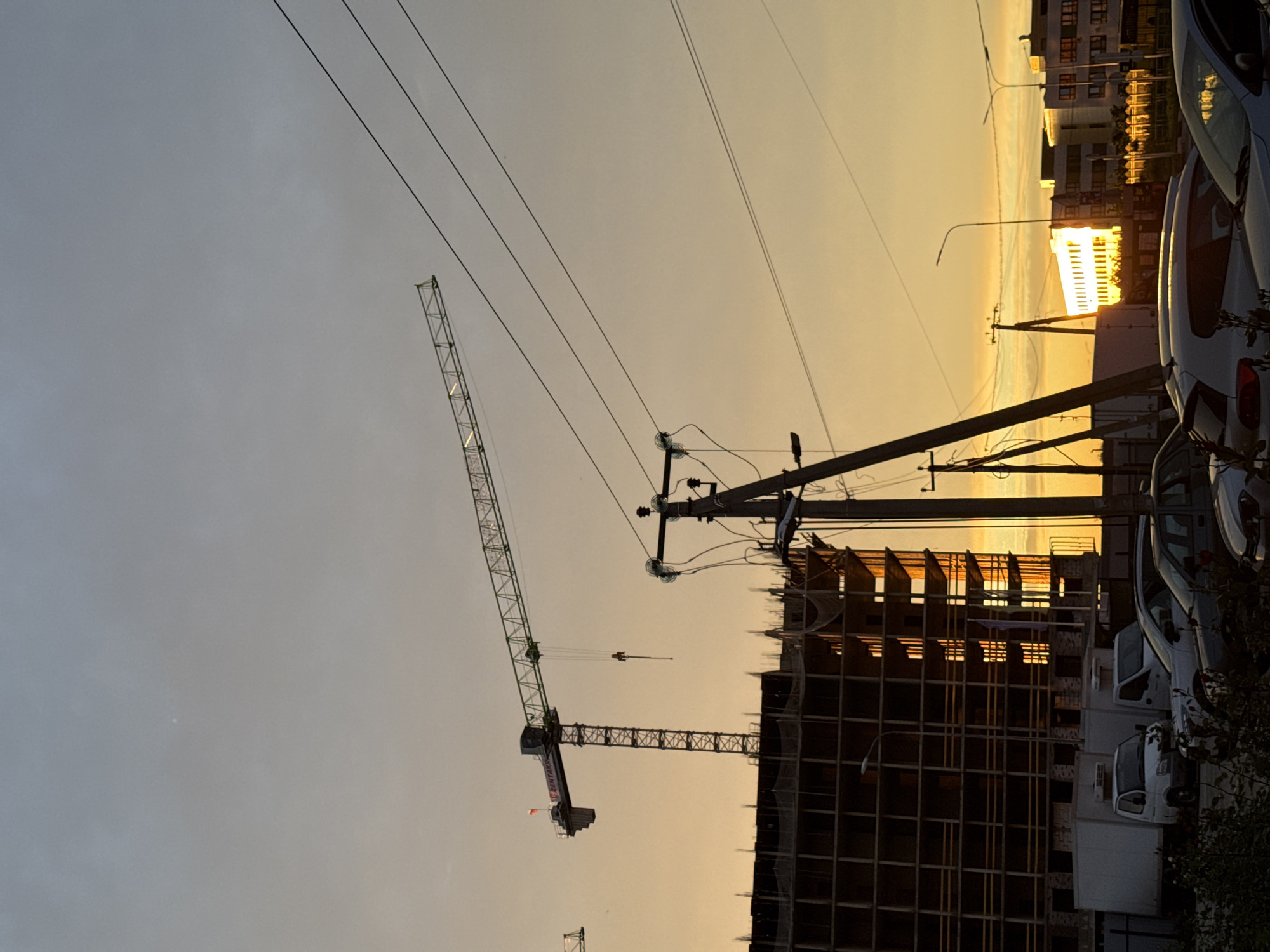 The image displays an urban or semi-urban scene during either sunrise or sunset, characterized by warm, golden light emanating from a low sun on the horizon. The sky exhibits a gradient, transitioning from a hazy light gray-blue at the top to vibrant orange and yellow hues towards the bottom right.
The primary subjects include a prominent construction site featuring a tall tower crane, likely a luffing jib type, with its lattice structure silhouetted against the sky. The brand "KENTASSY" is visible in red text on the crane's jib. Adjacent to the crane, an incomplete multi-story building is under construction, its skeletal frame of concrete or steel beams catching the golden light on its upper levels, while lower sections appear darker.
A dense network of overhead power lines crisscrosses the sky, anchored by several utility poles throughout the middle ground. In the right foreground and mid-ground, multiple cars, including white sedans or vans, are parked, their surfaces reflecting the warm, ambient light. Further in the background, additional buildings are visible, also illuminated by the low sun. The overall scene suggests an area undergoing development, captured during the "golden hour," creating strong contrasts and silhouettes against the luminous sky. No people are discernible in the image.