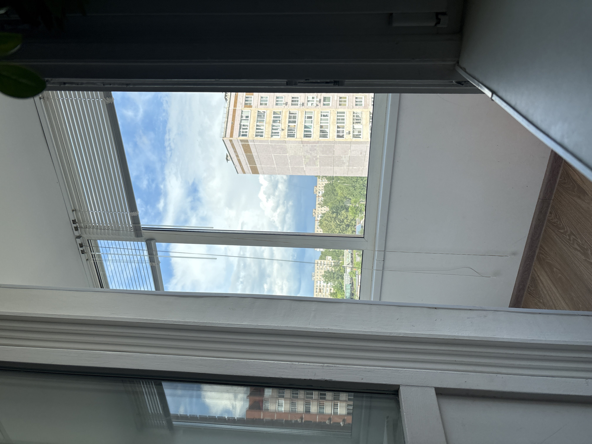 The image captures a view from inside a building in what appears to be Chaikovsky, Russia. The view is directed outwards through a window with partially drawn blinds, framing a tall, multi-story building across the street, with other residential buildings and greenery visible in the distance under a cloudy, blue sky. The window is part of an interior that includes white walls and light wood flooring. A portion of the window frame is visible, and there is a plant's foliage on the left side. The reflection on the window pane reveals a reversed image of the building seen outside.