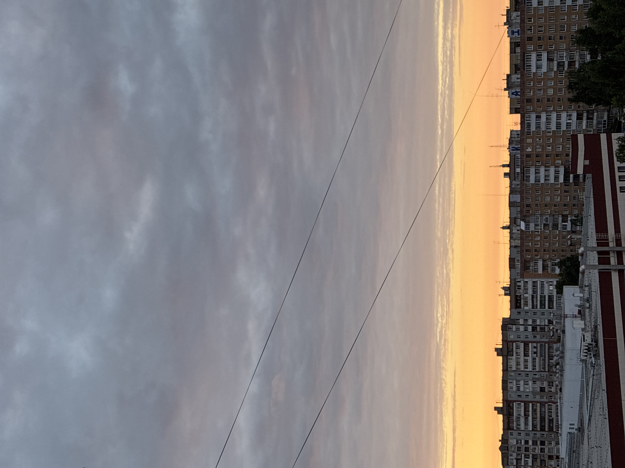 The image depicts an urban landscape during either sunrise or sunset, characterized by a vibrant orange and yellow glow at the horizon. The upper two-thirds of the sky are filled with a dense, textured layer of gray and light gray clouds, with a subtle pinkish tint near the horizon blending into the orange. Several dark, thin wires diagonally traverse the upper mid-section of the frame, running from the upper left towards the mid-right.
Along the horizon, a continuous line of multi-story residential apartment buildings is silhouetted against the bright sky. These buildings feature numerous uniformly shaped windows and balconies. On the rooftops of several of these structures, large dark Cyrillic letters are prominently displayed: "К" (K) on the left building, "А" (A) on a central building, and "И" (I) on a building to the right. Various antennae and small structures are also visible on the rooftops.
In the lower portion of the image, below the residential blocks, are several lower-profile industrial or commercial buildings with flat rooftops. One prominent building in the immediate foreground features a reddish-brown base and a white upper section, also with a flat roof equipped with ventilation units and pipes. Scattered trees are visible between these foreground structures and the apartment buildings. No people are discernible in the scene. The location is in the city of Unknown, Unknown.