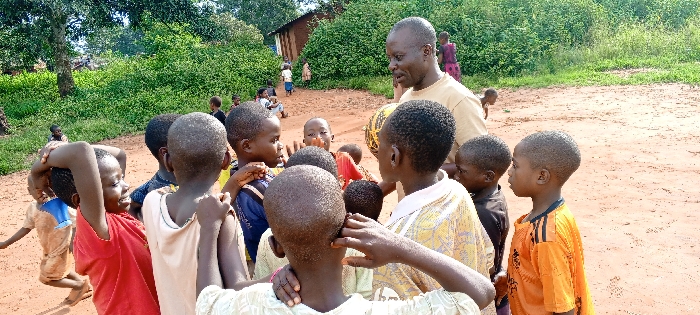The image depicts an outdoor gathering in Ngalula, Democratic Republic of the Congo, under bright daylight. A group of approximately ten children, ranging from early childhood to pre-adolescence, are clustered on reddish-brown earth. They are arranged in a loose semi-circle around an adult male positioned in the upper right quadrant of the frame. The adult, with short dark hair and stubble, wears a light-colored, short-sleeved t-shirt and holds a yellow and black patterned spherical object, consistent with a deflated ball, in his left hand. His gaze is directed downwards towards the children, suggesting engagement.
Among the children, one on the left in a red t-shirt smiles, with both hands near their head. Another child in an orange and black striped t-shirt is visible on the far right, facing left. In the immediate foreground, a child wearing a light-colored, patterned shirt has a hand resting on another child's shoulder. Other children are present, dressed in various casual clothing, including blue and black shirts. In the background, dense green foliage, encompassing trees and bushes, is prominent. Behind this vegetation, on the left, several simple, single-story structures with dark walls and lighter roofs are visible. Additional individuals, including children, are partially discernible in the distant background near these structures. The ground throughout the scene is uniformly composed of reddish-brown soil.