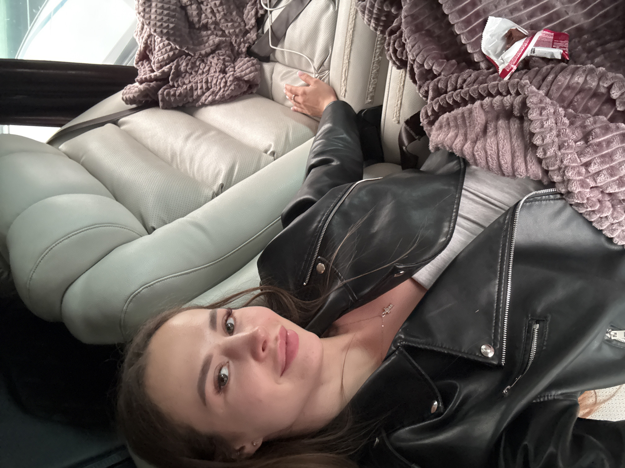 A young woman is lying down in a vehicle, possibly a car or van, in an unknown location in the city of Bielany Wrocławskie, Poland. She is wearing a black leather jacket and a light-colored top. She has brown hair and is looking directly at the camera with a slight smile. The interior of the vehicle features light-colored, quilted leather seating. There is a lavender-colored, waffle-textured blanket draped on the seat behind her. On top of the blanket, there appears to be an open package of a chocolate bar.