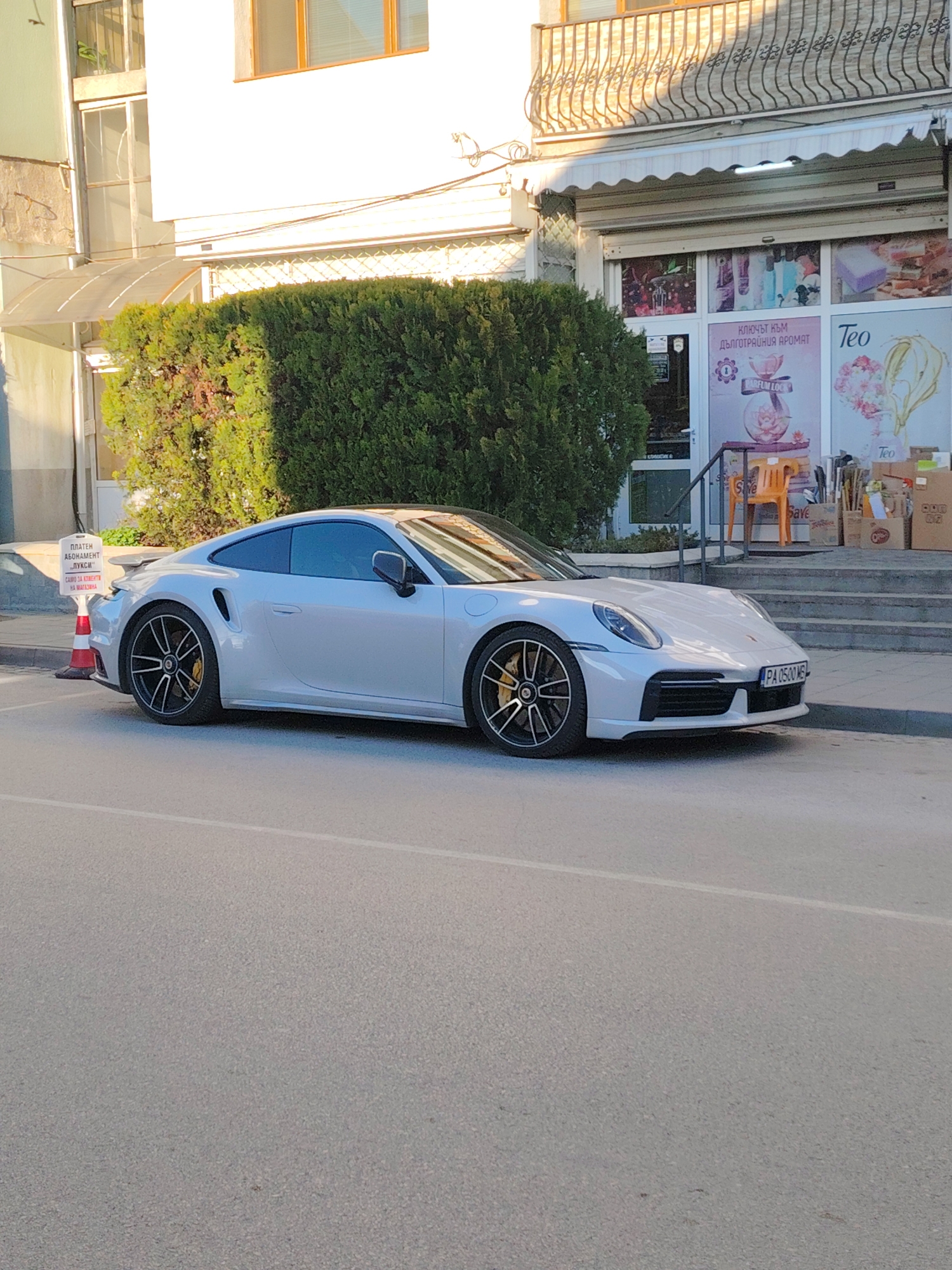 The image captures a white Porsche 911 Turbo S parked on the side of a street in Panagyurishte, Bulgaria. The car is parked in front of a storefront with an awning, a balcony above, and signage for a perfume shop and another business called "Teo". The shop's window displays perfume advertisements and related imagery. A sign next to the car indicates that parking is only for store customers. An orange cone stands next to the sign. The street is paved and occupies the majority of the foreground. The environment includes other buildings and greenery in the background, contributing to the urban landscape. The day appears sunny, casting shadows on the building and the street.